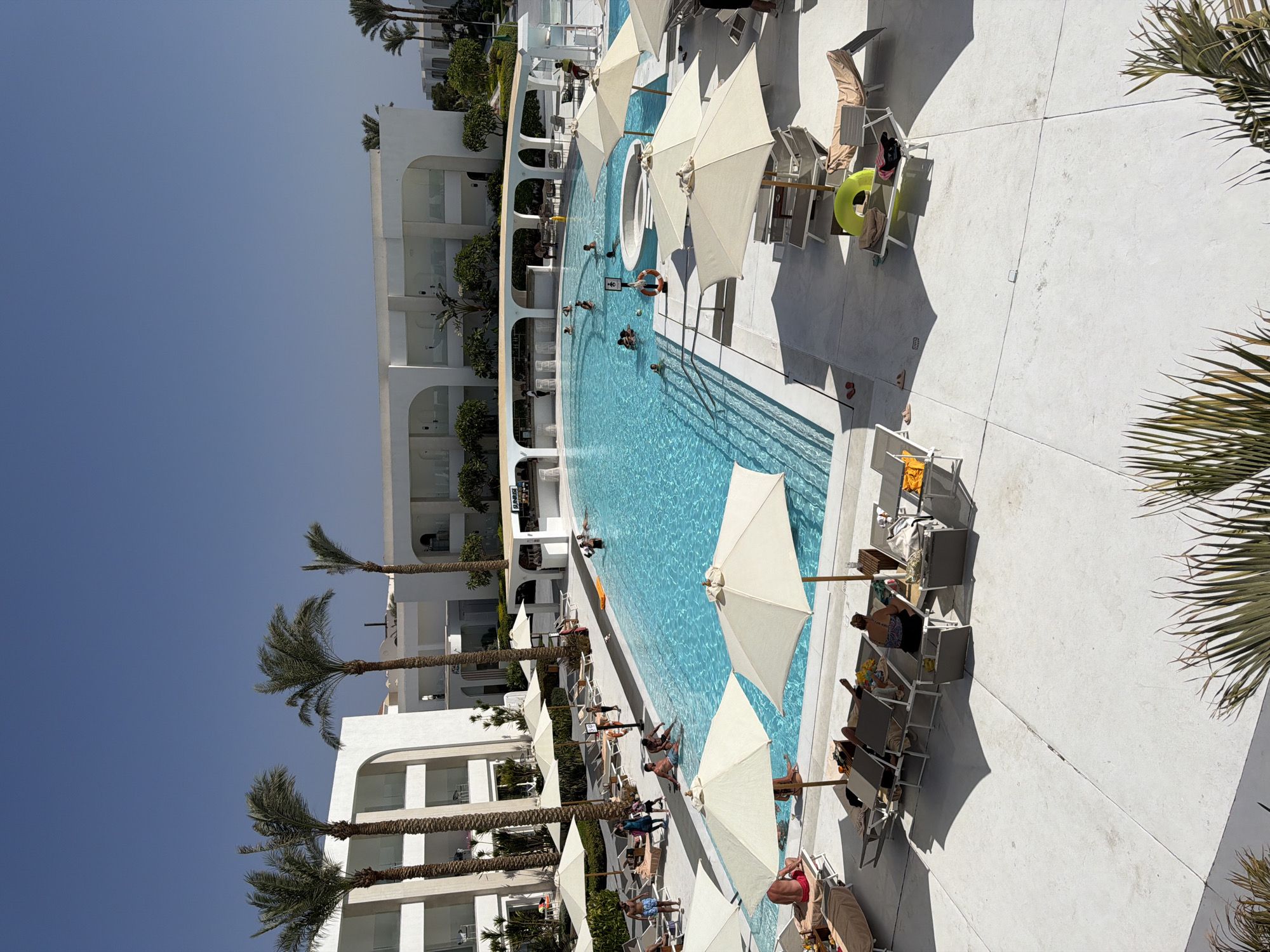 A high-angle, eye-level view captures an outdoor swimming pool area at a resort, likely in Sharm El Sheikh, Egypt, under a clear blue sky. The resort features modern white architecture with curved elements and palm trees scattered throughout.
The main focus is a large, irregularly shaped swimming pool filled with bright blue water. Several people are in the pool, some swimming, others socializing. There is a swim-up bar with people seated at it. Around the pool, white tiled paving surrounds lounge chairs and umbrellas. Many lounge chairs are occupied by sunbathers, some reading, others relaxing. There are several large, cream-colored umbrellas providing shade. Other items visible include towels, bags, a yellow inflatable ring, and a life preserver.
In the background, multi-story white resort buildings with balconies are visible, along with more palm trees. The overall atmosphere suggests a warm, sunny day at a vacation resort.