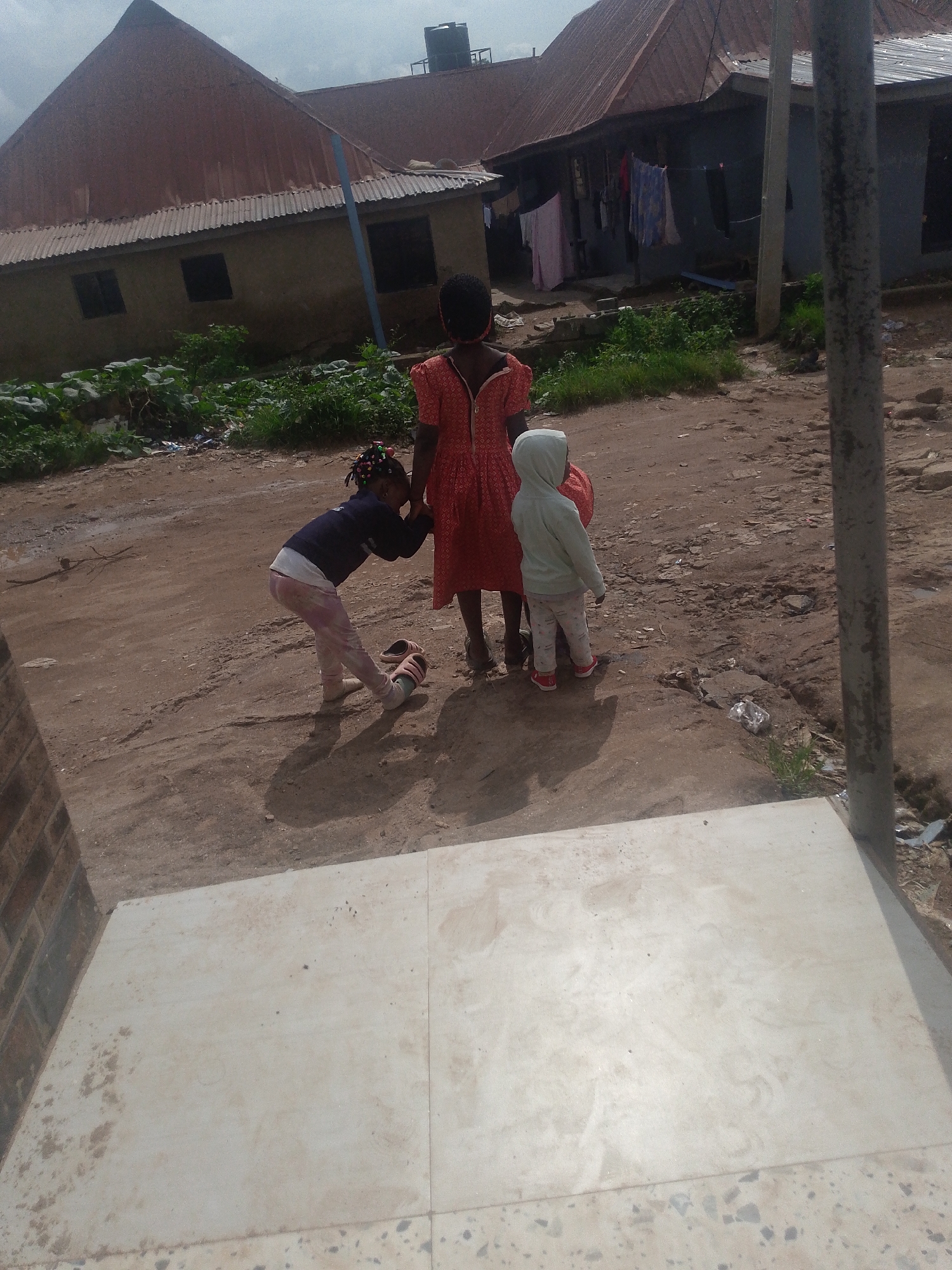 In Kurunduma, Nigeria, three children are pictured outdoors on a dirt road in front of a building with a corrugated metal roof. The child on the left, a young girl with braided hair, wears a dark blue long-sleeved shirt and light pink patterned pants. She is bent over, interacting with something on the ground or possibly a toy. To her right, a taller child in a red dress stands facing away from the camera. Beside the taller child, to the right, a third child in a light grey hooded sweatshirt and white patterned pants is also facing away. Clothing hangs on a line in front of the building. The environment appears somewhat unkempt, with scattered debris visible on the ground. The lighting suggests it is daytime, possibly overcast.