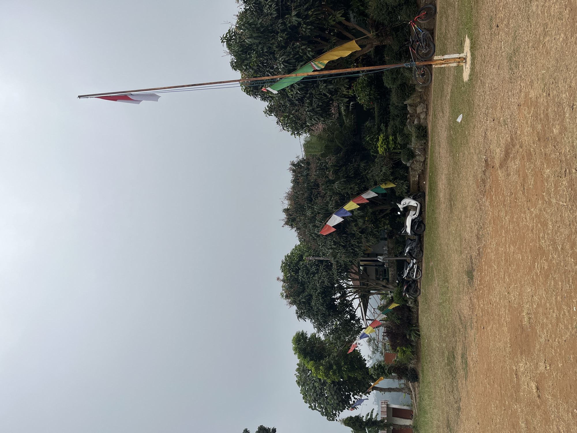 The image depicts an outdoor scene with a flag pole in the foreground, bearing the Indonesian flag. Behind the flag pole, there is a grassy field that transitions into a dirt path. In the mid-ground, several trees and shrubs are visible, some with colorful flags strung between them. A building with a dark roof is partially obscured by the foliage. To the right of the building, two bicycles are parked. Further to the left, three scooters are lined up. The sky is overcast and a hazy, light gray. The overall environment appears to be a rural or semi-rural setting. Without additional context, it is not possible to definitively place this scene within the city of Gembong, Indonesia.