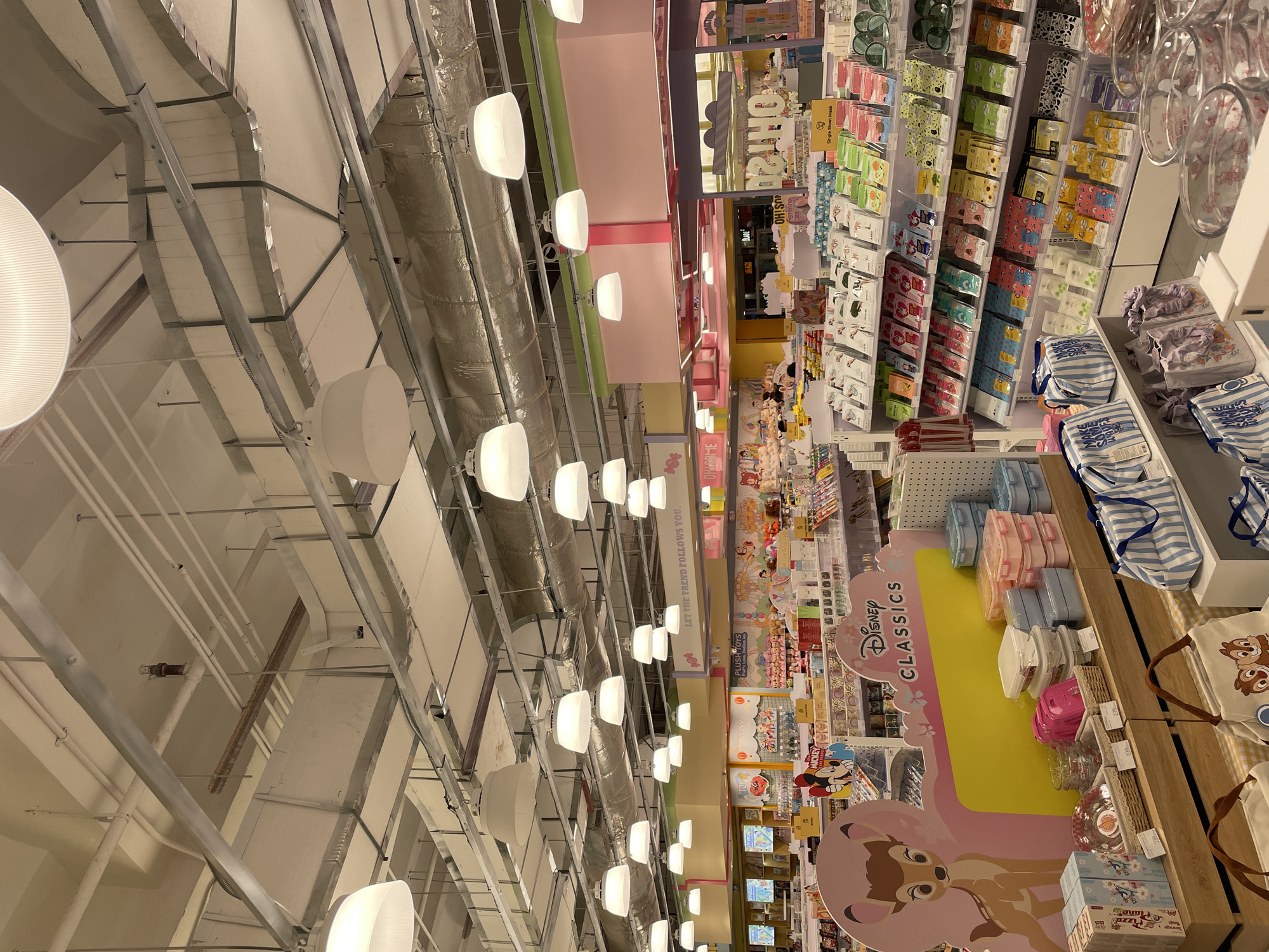 The image shows the interior of a retail store, likely a gift shop or a department store, with a focus on Disney merchandise. The ceiling is a complex network of exposed pipes, ducts, and light fixtures, suggesting an industrial or modern design. Rows of shelves are stocked with various products, including what appear to be facial masks, bags, and kitchenware. Prominently displayed is a section featuring "Disney Classics" with an image of Bambi. Further back, shelves are filled with plush toys and other Disney-themed items, including Mickey Mouse merchandise. The lighting is bright and evenly distributed, illuminating the merchandise effectively. The overall impression is of a well-organized retail environment catering to fans of Disney characters and related products. Based on available information, this store is located in SM Lanang Premier, Davao City, Philippines, a large shopping mall in the region. There are no people clearly visible in the scene.