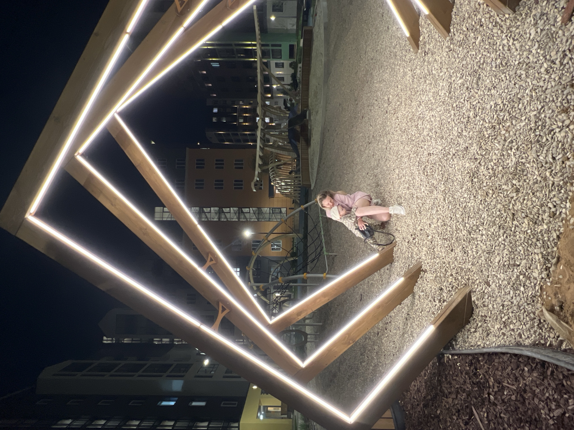 At night in Pervomaysky, Russia, a child in a pink outfit sits on a ground covered with small pebbles, next to a geometric wooden structure illuminated with white lights. In the background, buildings of varying heights and architectural styles are visible, some with lit windows. A playground structure is also partially visible behind the wooden frame. The scene is lit by the wooden structure lights and streetlights, creating a contrast between the foreground and the darker background.