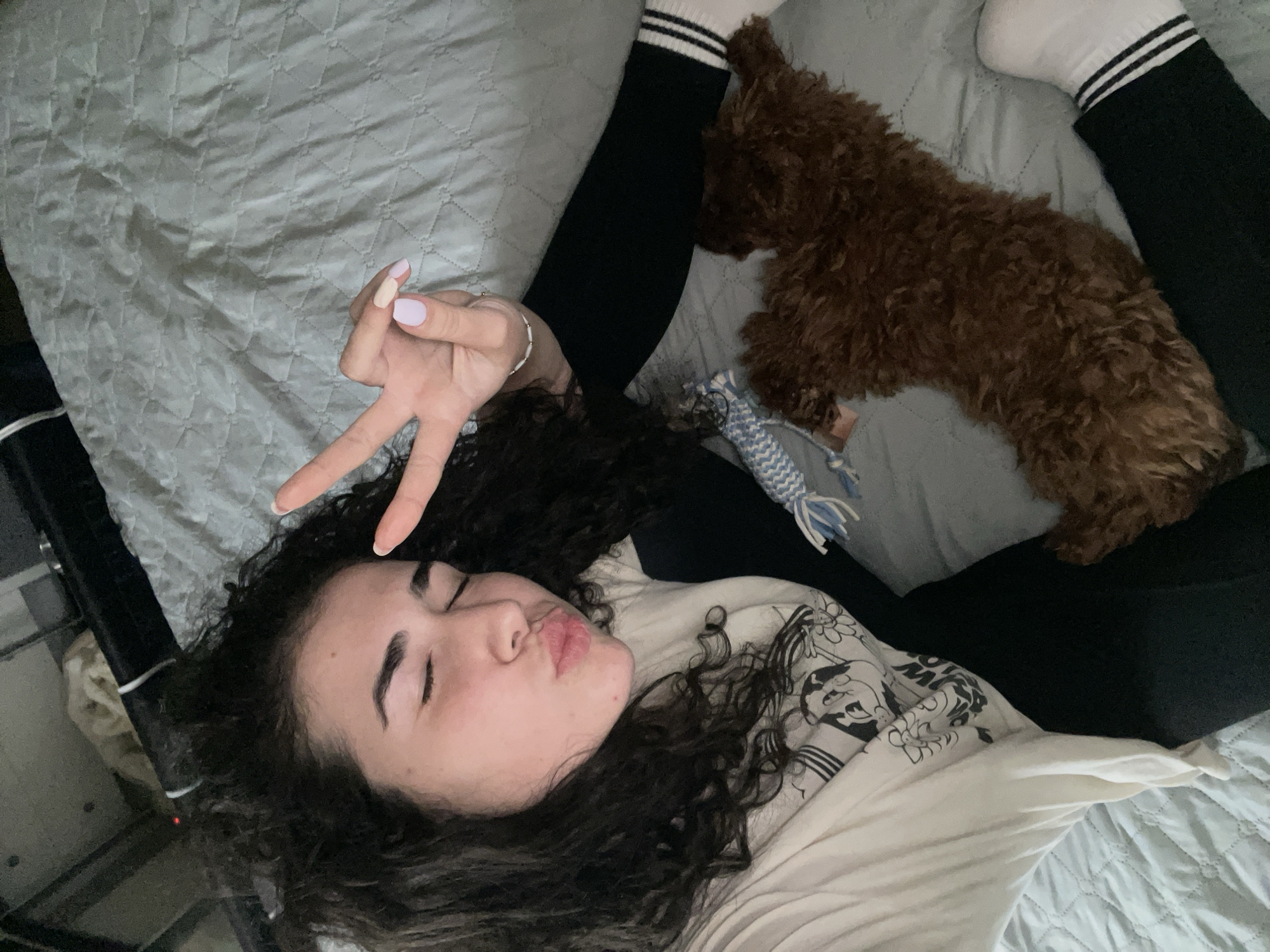 A young woman and her small, brown, curly-haired dog are captured in a high-angle shot, likely a selfie, while lying on a bed with light grey quilted bedding. The woman, with dark curly hair, lies on her back with her head turned slightly to her right. Her eyes are closed, and her lips are playfully pursed. She raises her left hand, making a peace sign, revealing light pink nail polish and a thin bracelet. She wears a light-colored long-sleeved shirt with a black graphic design featuring abstract shapes and possibly cartoon-like elements, along with dark leggings and white socks with two black stripes. Her dog, with rich brown, curly fur, rests calmly near her legs on the right side of the frame. A blue and white braided dog toy is visible near the dog's head. The scene conveys a relaxed, casual moment indoors, illuminated by soft, ambient lighting. No explicit text is clearly legible on her shirt, and no specific location cues for Sofia, Bulgaria are visible within the image.