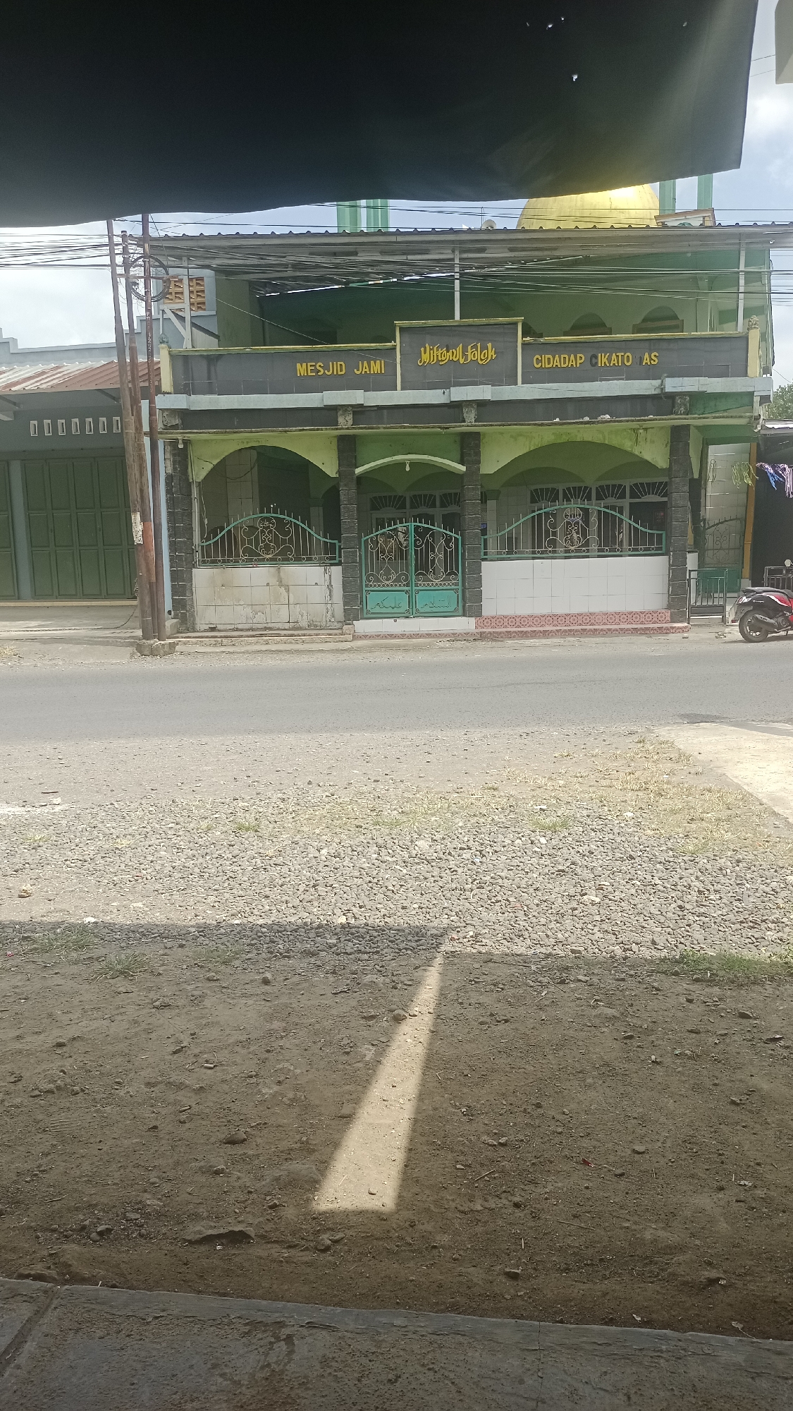 The image captures a street view of a building with a golden dome, identified by signage as "MESJID JAMI HIDAYAT FOLK CIDADAP CIKATOMAS". The building appears to be a mosque, painted in shades of green and gray. It features arched entrances, ornate green metal gates, and tiled walls. In front of the mosque, a paved road separates it from a gravelly area and a dirt patch where a shadow is cast. To the left of the mosque, a green-gated building with corrugated metal roofing is visible. A red motorcycle is parked to the right of the mosque. The overall environment suggests a public street in a potentially tropical or subtropical climate, with utility poles and overhead wires present. The image is taken from a low angle, looking slightly upwards towards the mosque.