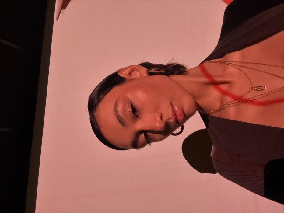 A woman with slicked-back hair and hoop earrings poses at an angle. Her skin is glowing under a warm light. She wears layered gold necklaces, one with a small pendant. A red, angular design is painted on her neck and chest. She is wearing a dark top. Part of a door frame is visible on the left side of the frame and the background is light pink.