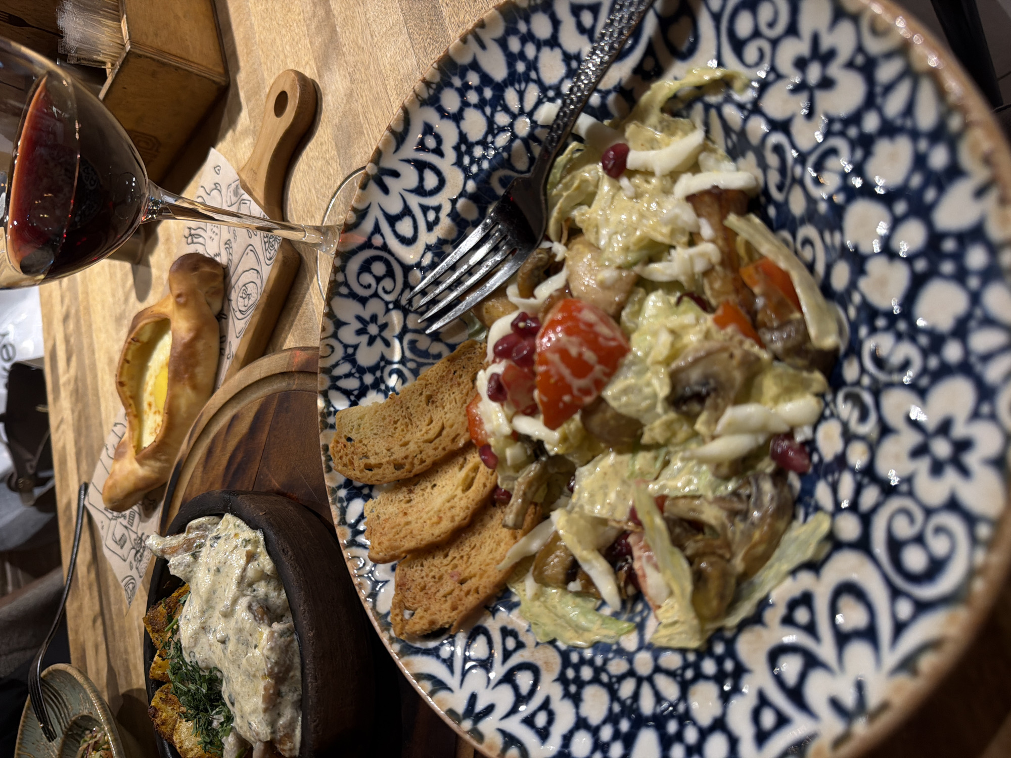 The image depicts a table setting with a partially filled wine glass, a boat-shaped bread, and two dishes. One dish is a blue and white patterned plate containing a salad with sliced bread. The salad consists of lettuce, mushrooms, tomatoes, pomegranate seeds, and a creamy dressing. A black fork is placed on the plate. The second dish appears to be a stew with a creamy sauce and greenery, served in a dark, possibly wooden bowl. All are situated on a wooden table or surface. The location is Izhevsk, Russia.