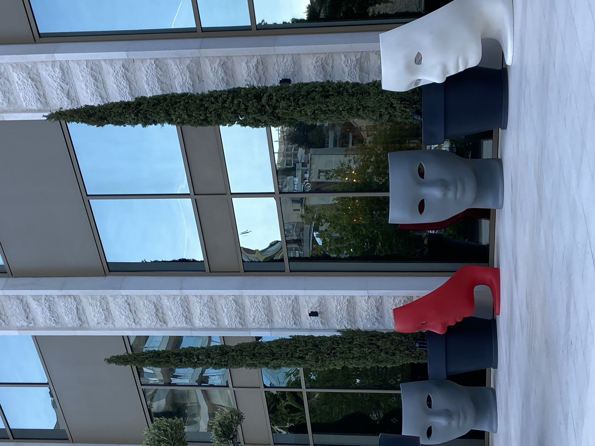 The image shows an outdoor display of three sculptures placed on a light-colored surface, likely a sidewalk or patio. Each sculpture consists of a head-shaped mask in a different color (white, gray, and red), each positioned atop a black planter or pedestal. In the background is a multi-story building with large rectangular windows reflecting the sky and surrounding greenery, typical of an urban landscape. Columnar evergreen trees are planted near the base of the building. The overall scene conveys an artistic or decorative arrangement in an urban environment, with the city of Budva, Montenegro, as the probable location.