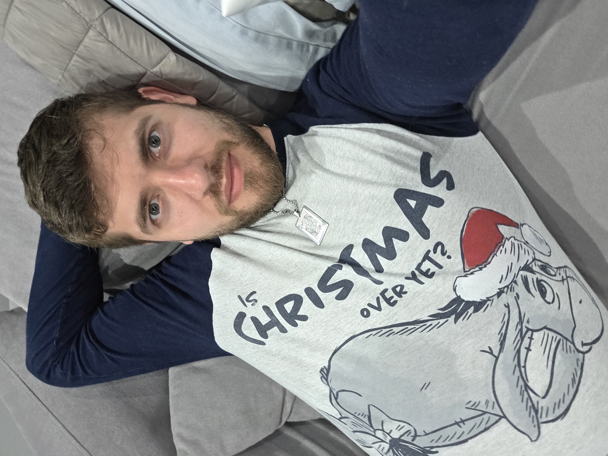 A man lies on a gray couch with his hands behind his head. He is fair-skinned with short brown hair and a beard. He is wearing a gray and navy blue long-sleeved shirt with the words "Is Christmas Over Yet?" and a cartoon image of Eeyore wearing a Santa hat. He also wears a silver necklace with a rectangular pendant. The location is unknown and there is no indication that the image was taken in Yagodovo, Bulgaria.