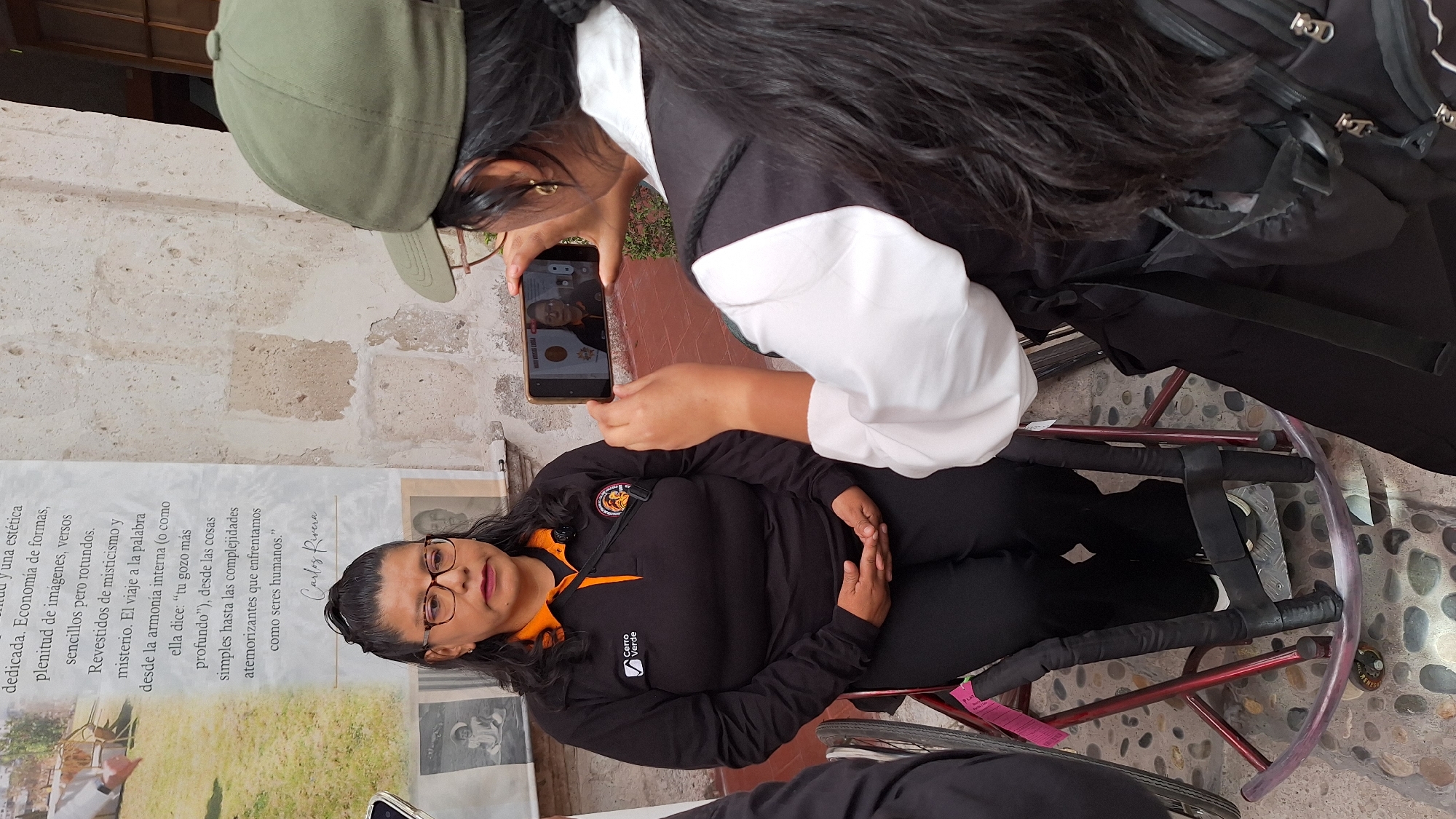 Two women are captured in a daytime scene, likely outdoors or in a semi-enclosed area with stone walls and a textured stone ground. One woman, wearing a green baseball cap and a black and white top, is holding a black smartphone, seemingly taking a photograph. The phone's screen clearly displays a close-up image of the other woman.
The second woman is seated in a red-framed wheelchair, wearing glasses, a black long-sleeved polo shirt with an orange collar, and dark trousers. Her shirt bears a "Cerro Verde" logo on the left side of her chest. She has dark hair and a neutral expression as she looks forward.
Behind the seated woman is a tall, vertical banner or poster. Visible text on the banner is in Spanish, including phrases like "dedicada. Economía de formas, plenitud de imágenes, versos sencillos pero rotundos," and a longer quote that discusses internal harmony, profound joy, simple things, and terrifying complexities faced by humans. The text is attributed to "Carlos Rivera." The image captures a moment of photography, possibly at an exhibition or event.