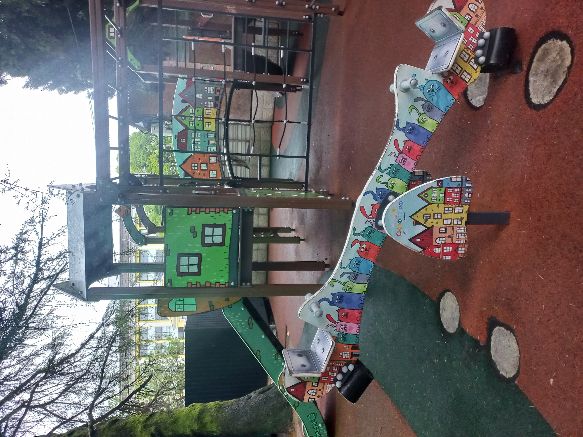 The image shows a playground in Sochi, Russia. There's a seesaw with colorful cat and house designs, along with other playground structures like climbing nets, slides, and a house-shaped structure. The ground is a soft, safety surface with green and brown colors. A tree is visible on the left side of the image. The sky is overcast.