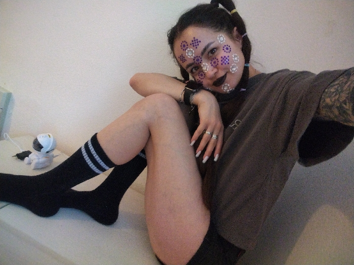 An indoor scene in the city of Noviy, Russia, displays a female subject seated or reclining against a plain, light-colored background. She directly faces the camera. Her dark hair is styled into two pigtails secured with yellow ties. Her face is adorned with dark lipstick and multiple adhesive decorative stickers, consisting of purple circular patterns with white dots and white circular patterns with reflective dots, applied to her cheeks and forehead. She wears a loose, dark grey t-shirt, revealing a full sleeve tattoo on her left arm. A dark watch or bracelet is visible on her left wrist, and her left hand features several rings on the fingers and white-tipped fingernails. Her legs are bent, with the left leg crossed over the right; the left foot is clad in a black sock with two white stripes. To her left, on the same light-colored surface, a small white spherical device with a black face and a white cable is present, next to a small, white, plush object.
