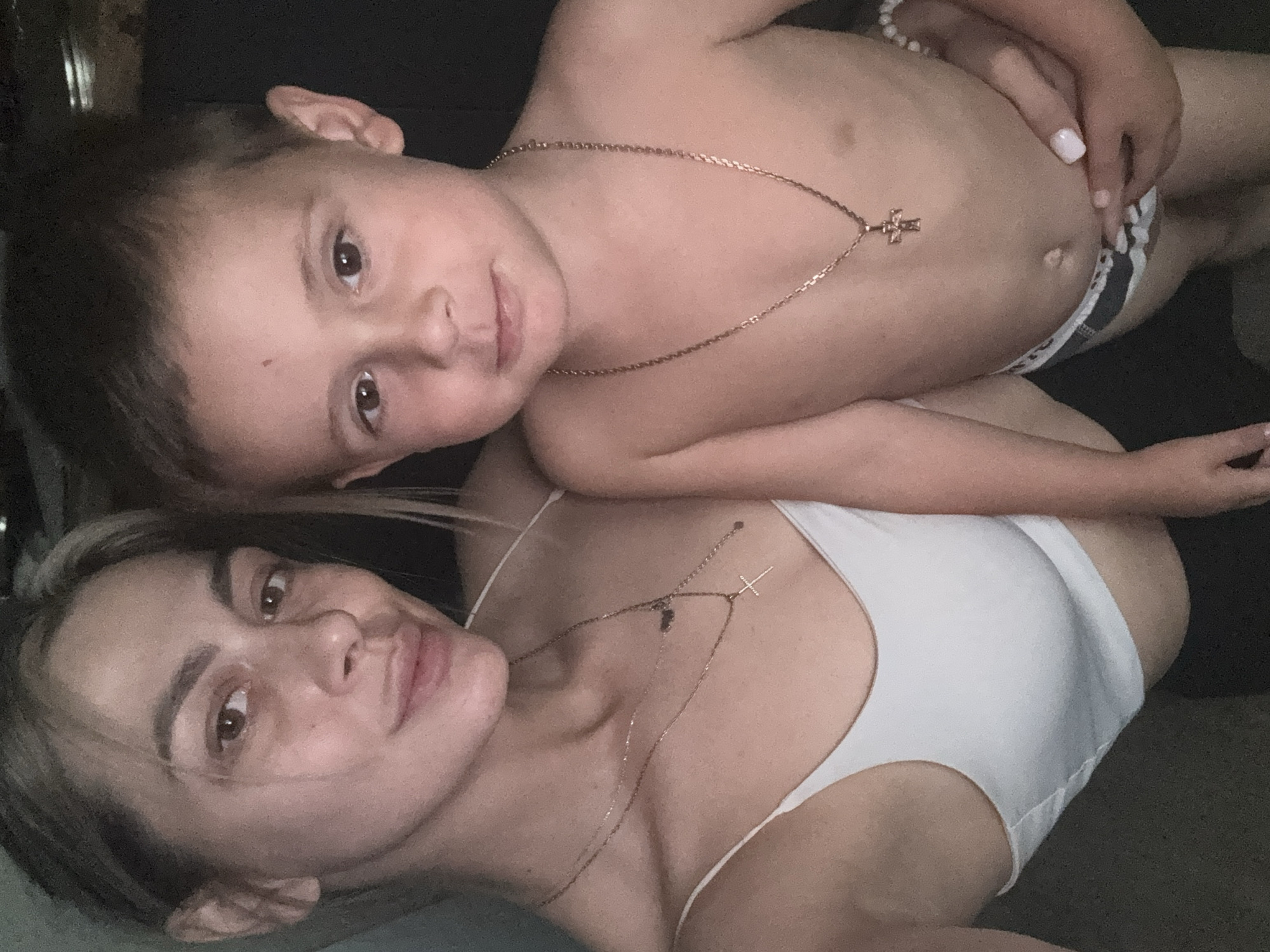 The image shows a close-up, vertically oriented selfie of a woman and a young boy. The woman is in the foreground, lying on her side and looking towards the camera with a soft smile. She has light skin, dark eyes, and dark hair that is partially visible. She is wearing a white bra. The young boy is positioned on top of her, also lying on his side and looking towards the camera. He has light skin, brown eyes, and dark hair. He appears to be topless, wearing dark and white patterned underwear. Both individuals are wearing gold necklaces with cross pendants. The background is dark and appears to be an indoor setting, possibly a bed or sofa. The lighting is dim, suggesting it might be evening or indoors with artificial light. The overall mood seems intimate and relaxed, possibly capturing a moment of bonding between the mother and child. There is no visible text in the image. Given the provided location context of Tula, Russia, it is implied that this scene is taking place within that region.