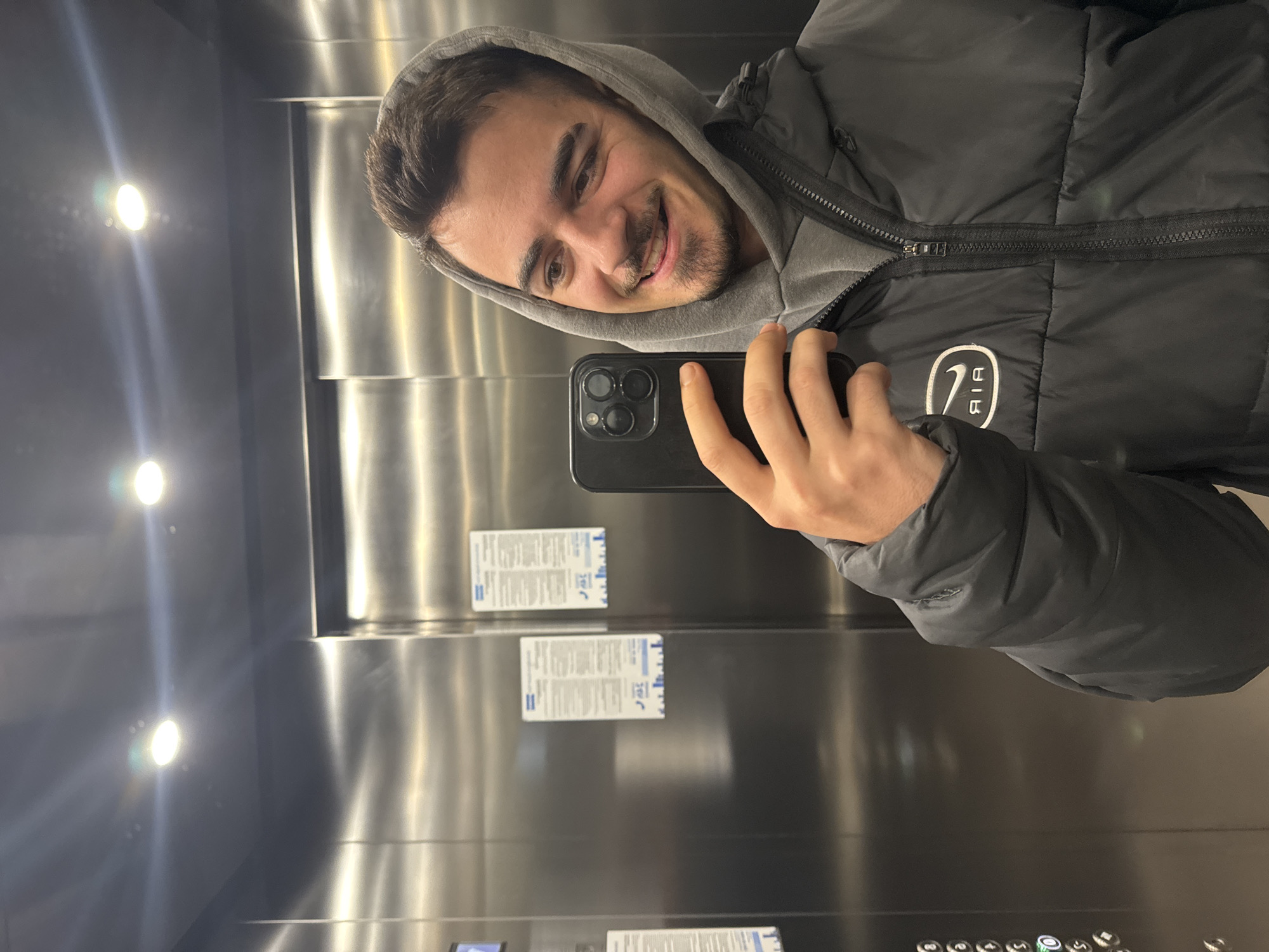 A man is taking a selfie in an elevator. He is wearing a grey hooded sweatshirt and a black puffy jacket with an "AIR" logo on the right breast. He's holding a black smartphone in his right hand. The elevator has brushed metal walls with various informational notices and a panel of buttons at the bottom. The location appears to be in a city, possibly Sofia, Bulgaria given the user context. The elevator is well-lit. The man is smiling at the camera.