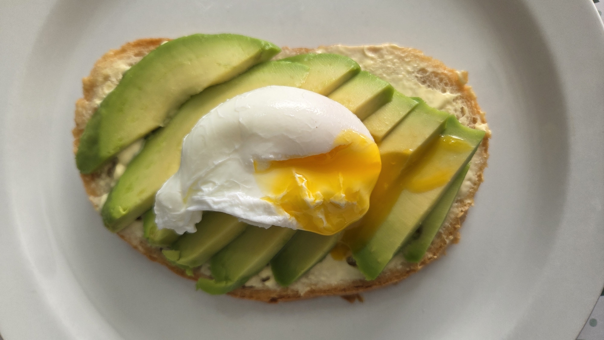 A close-up, overhead shot shows an avocado toast on a white plate. The toast has a slice of bread topped with a creamy spread, fanned slices of avocado, and a poached egg. The egg is perfectly cooked with a runny yolk that has spilled over the avocado. The background is a plain white plate, suggesting a simple meal setting. The image does not contain information about the location in Tynset, Norway.