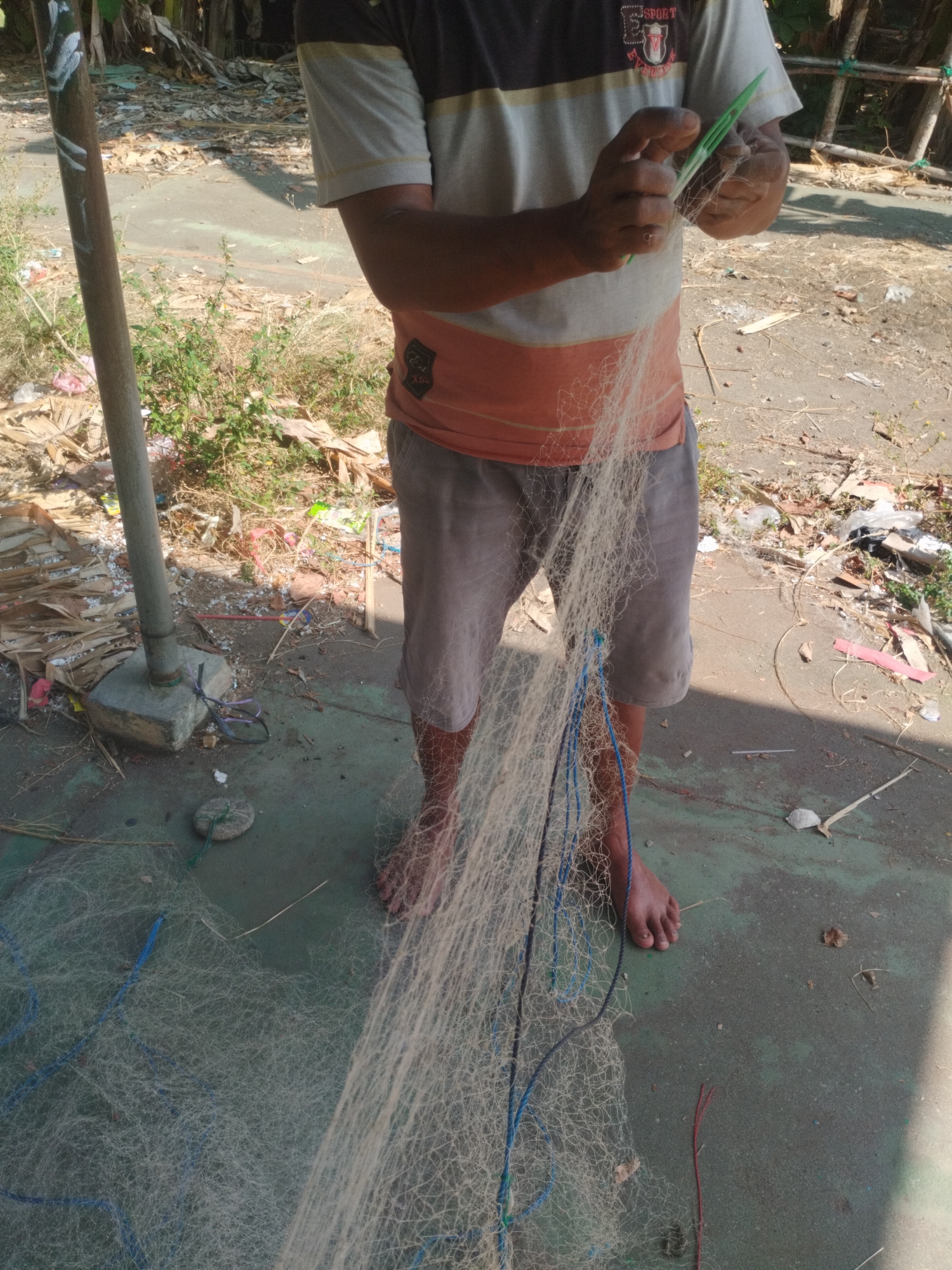 The image depicts a person in Kelurahan Cilacap, Kecamatan Cilacap, Indonesia, engaged in mending a fishing net. The individual, wearing a striped t-shirt and shorts, is shown holding the net and a green tool, likely a netting needle or shuttle, to repair it. The net is spread out on the ground, which appears to be a rough, outdoor surface with some scattered debris, including leaves, plastic waste, and building materials. A metal pole with a concrete base is visible to the left, possibly part of a structure or fence. The background suggests an outdoor environment with some vegetation and foliage. The overall scene conveys a sense of everyday labor and the tools associated with fishing activities.