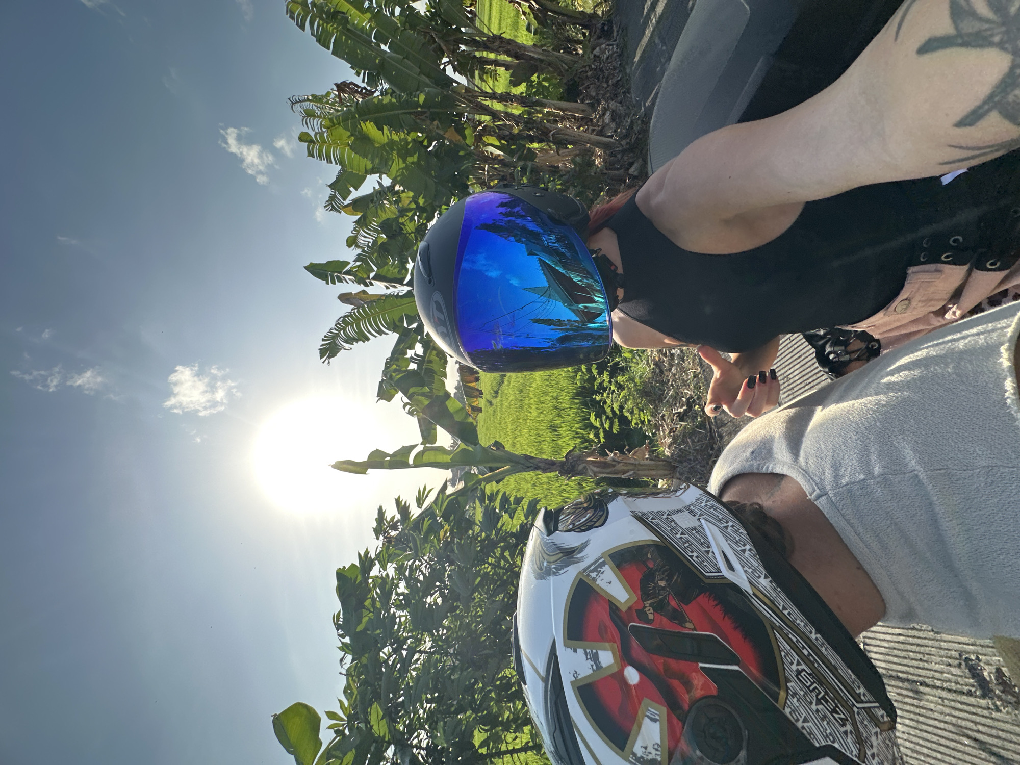 In North Kuta, Indonesia, two individuals wearing helmets are on a scooter. The person in the foreground on the left wears a white helmet with a red and black design. The person on the right wears a black helmet with a blue visor reflecting a building and trees. The background shows lush green rice fields, banana trees, and a bright sun in a clear sky with a few clouds. The road surface appears to be made of grey concrete with textured lines.