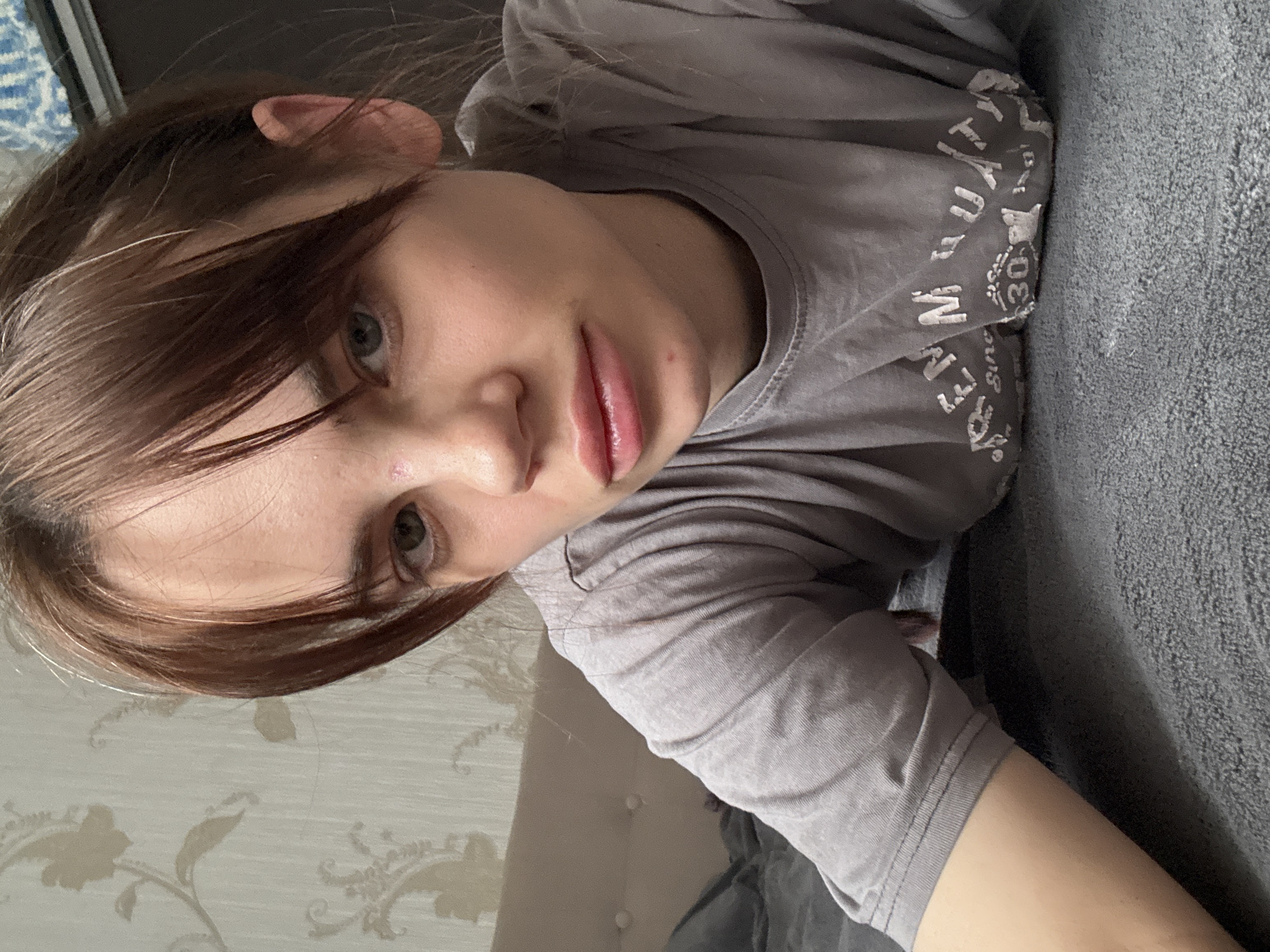 A young woman is lying on her side, resting on a gray upholstered bed or couch. She is wearing a gray t-shirt with white lettering on the upper right side. Her brown hair is parted on the side and falls across her forehead. She has light skin, green eyes, and full lips. The background includes a wallpapered wall with a floral pattern. The setting is a domestic interior. The image is rotated counterclockwise, and there is no evidence to support its location in Rostov-on-Don, Russia.