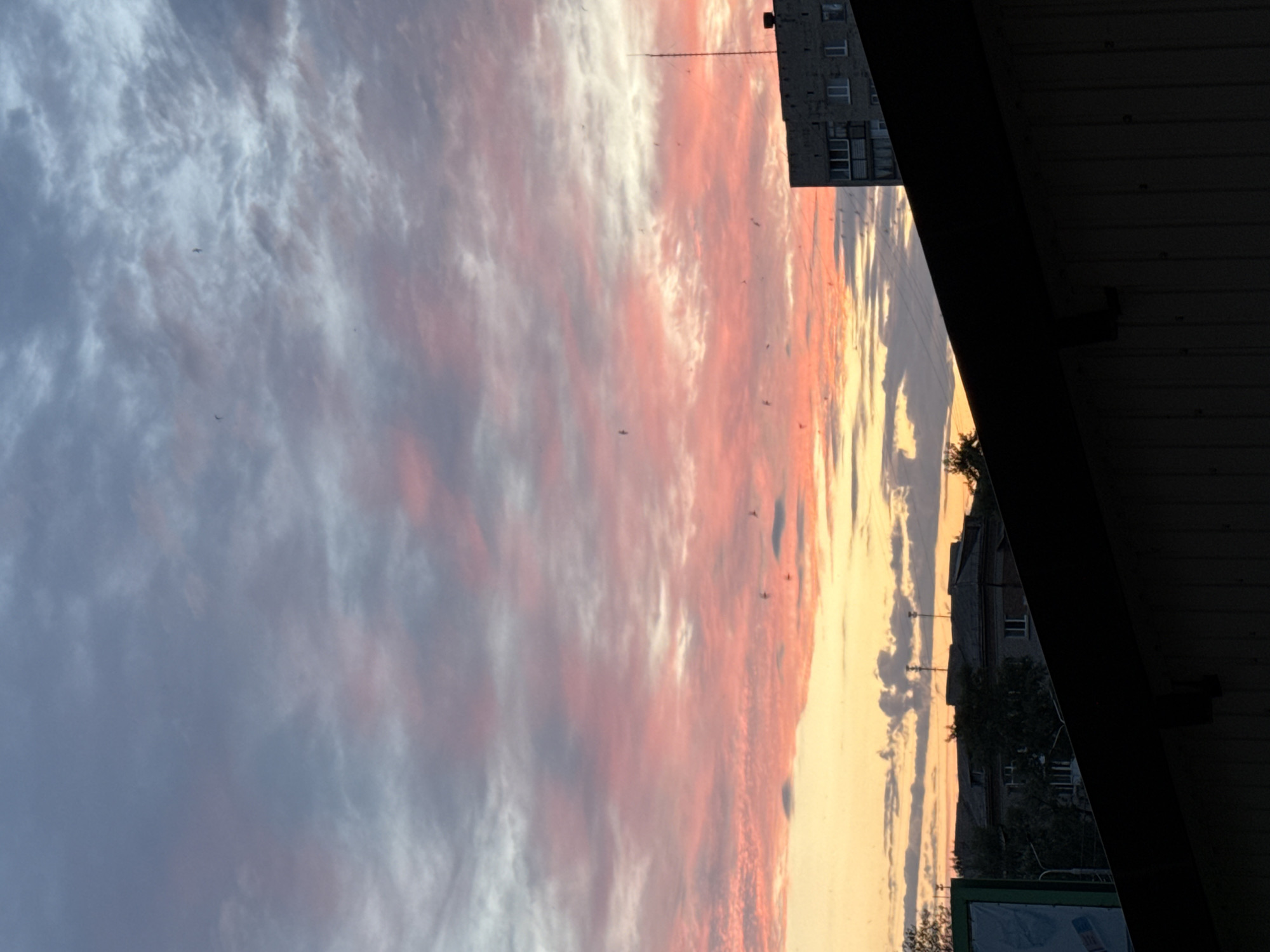 The image captures a sunset over the city of Luhansk, Ukraine. The sky is filled with dramatic clouds displaying hues of pink, orange, and gray, illuminated by the setting sun. Silhouetted against the vibrant sky are the rooftops of several buildings and a tall antenna. The foreground is dominated by the dark, angled roof of a structure, obscuring the lower portion of the scene. Some small, dark specks, possibly birds or dust, are visible scattered across the sky. The overall impression is one of a quiet urban evening, with the natural beauty of the sunset contrasting with the man-made structures below.