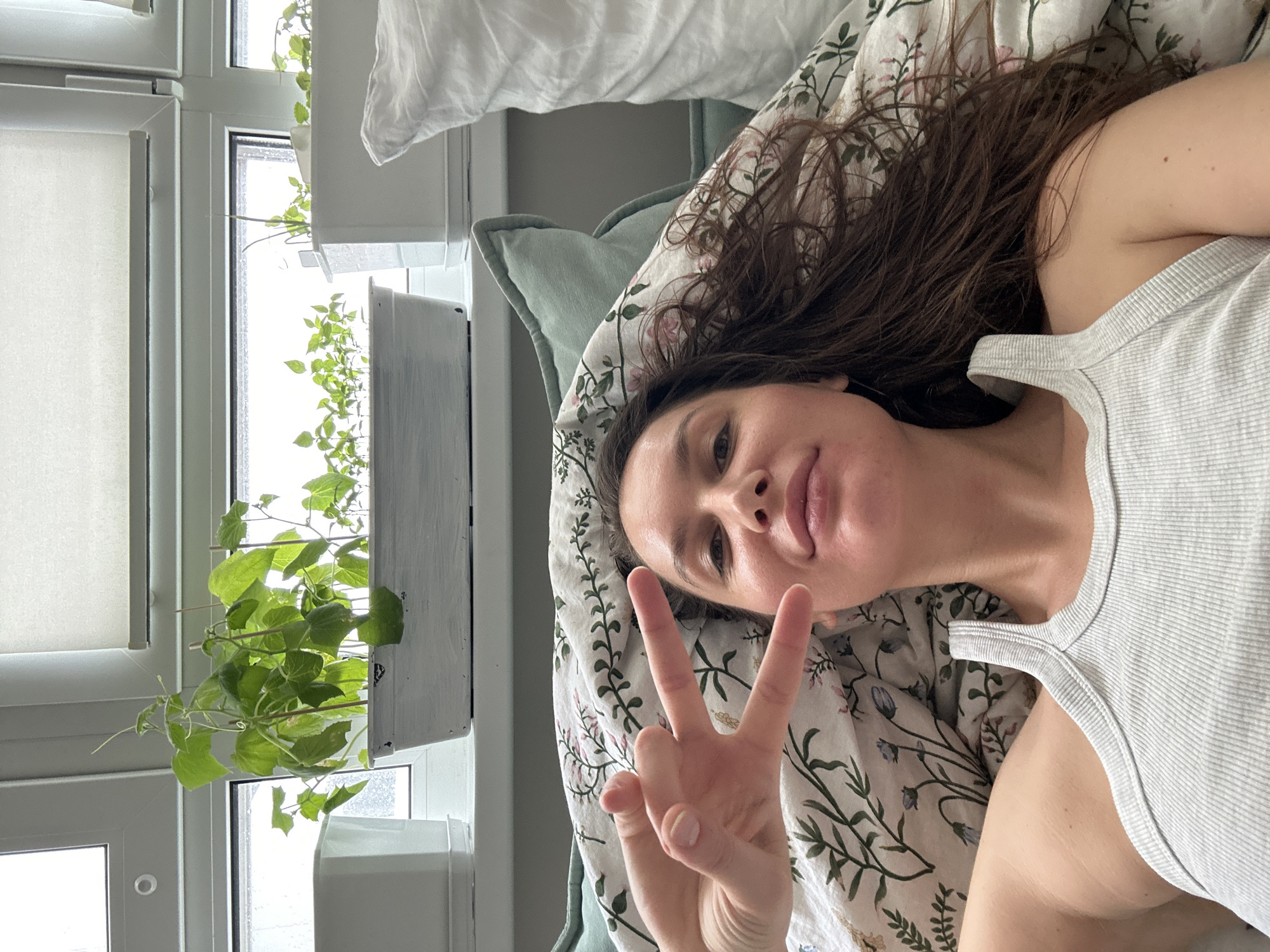A woman is lying on a bed, facing the camera, and giving a peace sign with her right hand. She has long, brown hair and is wearing a light-colored tank top. The bed has a floral-patterned sheet and a light green pillow. Next to the bed is a window with white frames and a white planter box containing green plants. The location is possibly Khimki, Russia.