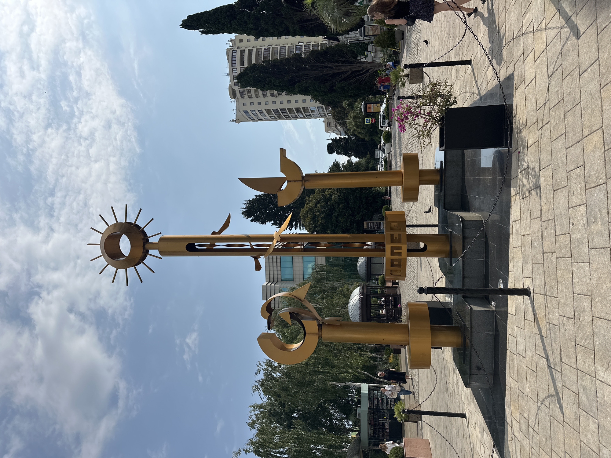 A golden abstract sculpture, possibly representing the sun, a bird, and a dolphin or a stylized "G," stands prominently in a paved public square. The sculpture is adorned with chains and bollards, suggesting it is a protected monument. In the background, people walk and shop at kiosks, while tall cypress trees and a multi-story building frame the scene. A bright blue sky with scattered clouds completes the urban landscape, which is identified as being in Sochi, Russia.