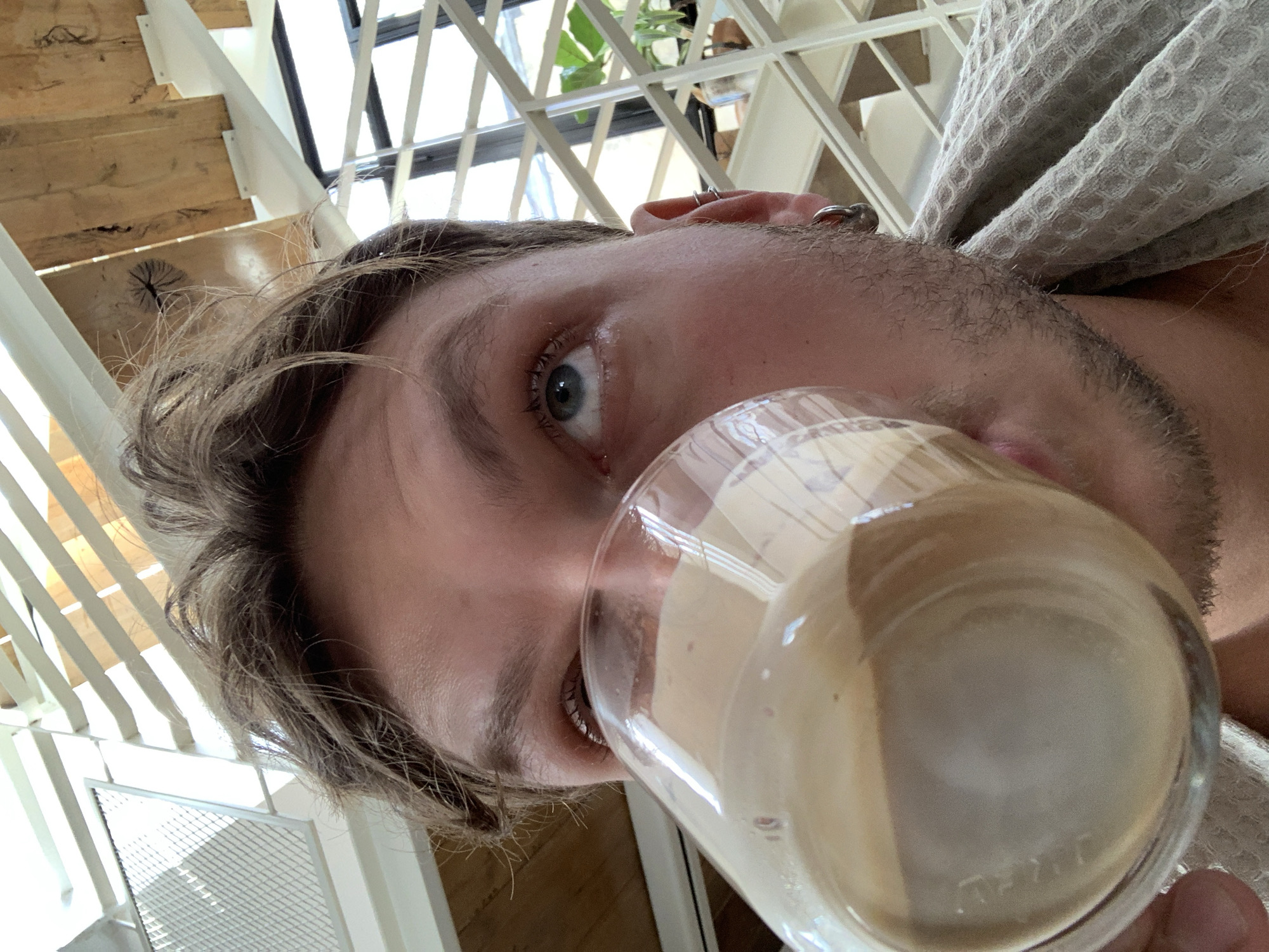 A close-up, tilted-angle shot shows a man drinking from a glass. He has fair skin, light brown hair, stubble on his face, and a blue eye visible. He wears a gray waffle-patterned robe or towel. A glass of a milky-beige liquid is held up to his mouth. The background is a bright interior with white walls, stairs with light wood treads, and a window or skylight with white latticework. The environment suggests a modern home or apartment. There is no information to ascertain the location of the subject is in Tel-Aviv.