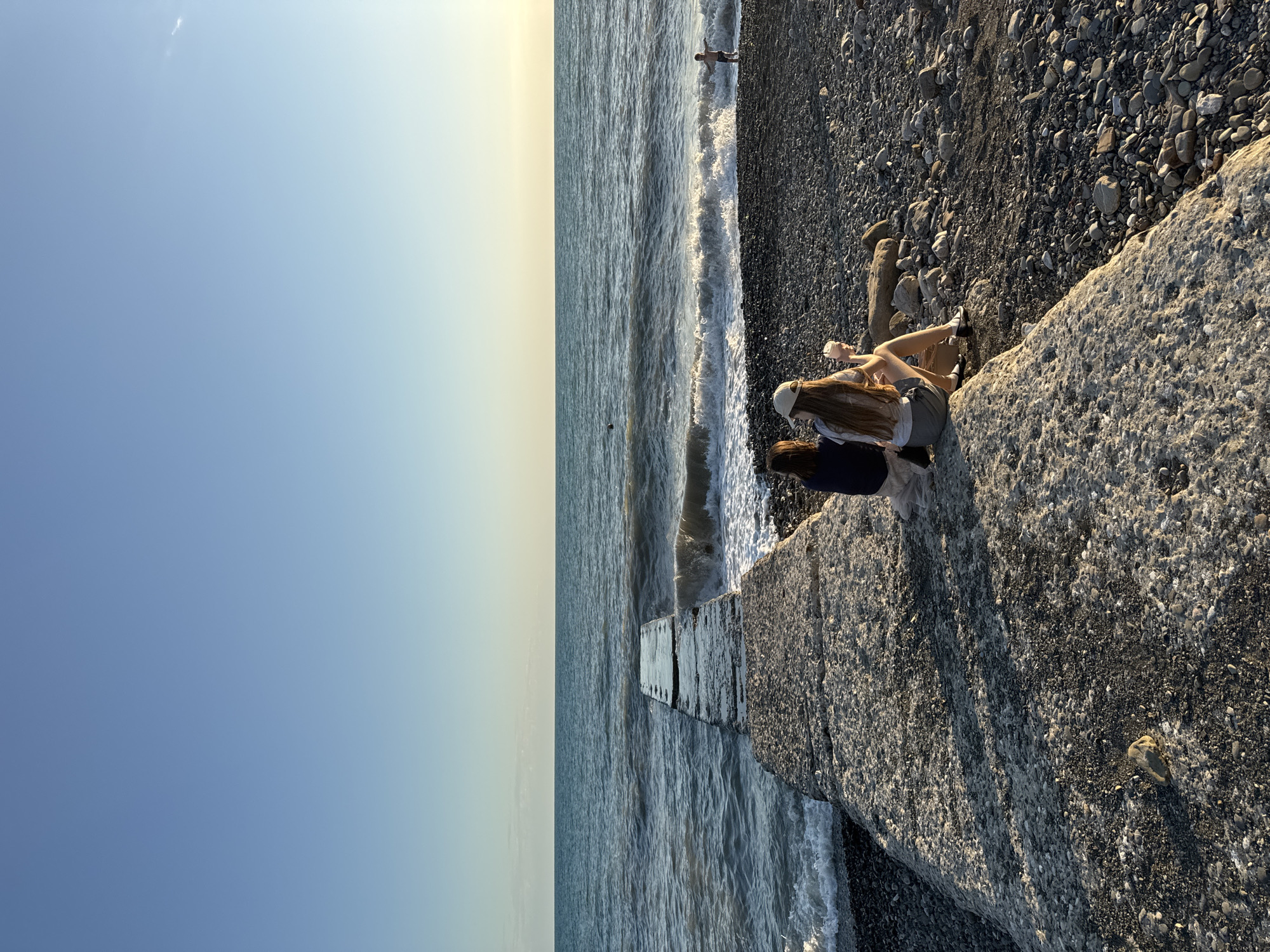 The image depicts two young women sitting on a large, rough-textured rock by the sea. The woman closer to the viewer has long, light brown hair and is wearing a white cap. She is facing away from the camera and appears to be looking out at the water. The second woman, partially obscured, is wearing a dark top and is sitting beside the first. Both are casually dressed, suggesting a relaxed atmosphere.
The scene is a pebbly beach with the sea stretching out towards the horizon. Waves are gently rolling onto the shore. A concrete structure, possibly a pier or breakwater, extends into the water from the right side of the frame. In the distance, a lone figure can be seen standing in the water.
The sky is a clear gradient of light blue to pale yellow, indicating it is either early morning or late afternoon, with the sun casting a warm glow. The overall mood is serene and peaceful. The location appears to be a coastal area, consistent with Sochi, Russia, known for its beaches and seaside resorts. There is no visible text in the image.