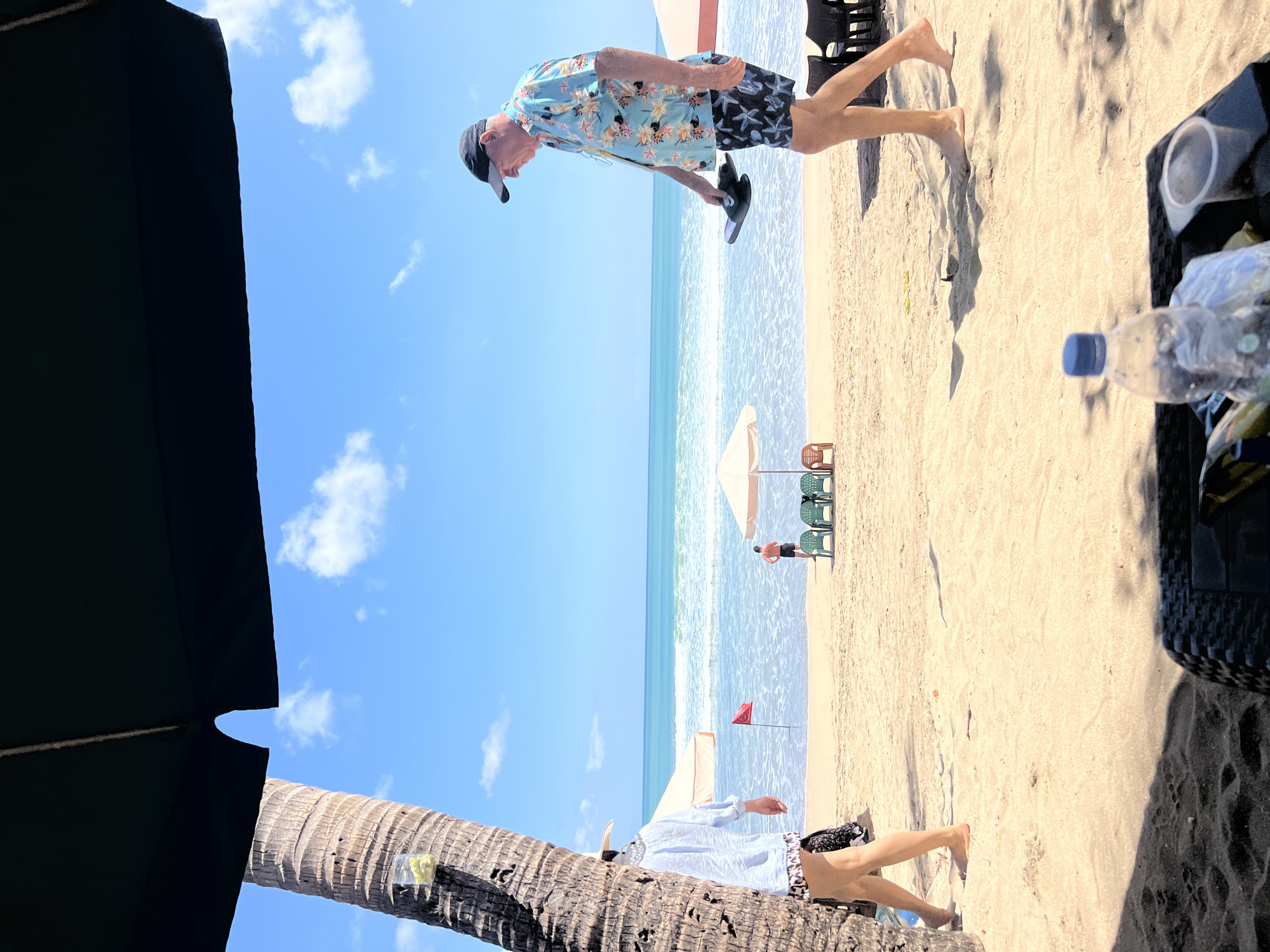 A tropical beach scene in Kuta, Indonesia, is depicted under a bright, sunny sky with scattered white clouds. The foreground is dominated by the dark shade of a large umbrella and a woven texture likely belonging to a table and chairs. A plastic water bottle with a blue cap and a clear plastic cup are visible on the woven surface. To the left, the trunk of a palm tree stands tall, with a small clear plastic container holding what appears to be lime wedges attached to it.
In the middle ground, the sandy beach stretches towards the ocean. Two people are walking on the beach. On the left, a person wearing a white shirt and patterned shorts walks away from the viewer. On the right, a man wearing a floral short-sleeved shirt, dark shorts with a white star pattern, and a black baseball cap walks towards the right, holding a pair of black flip-flops.
Further back, beach umbrellas and chairs are set up on the sand. A small red flag stands in the shallow water near the shoreline, and a lone figure is visible near a cluster of green chairs. The ocean waves are gently rolling onto the shore, and the horizon is marked by the clear blue sea meeting the bright sky. The overall atmosphere is one of a relaxed beach day in a tourist destination.