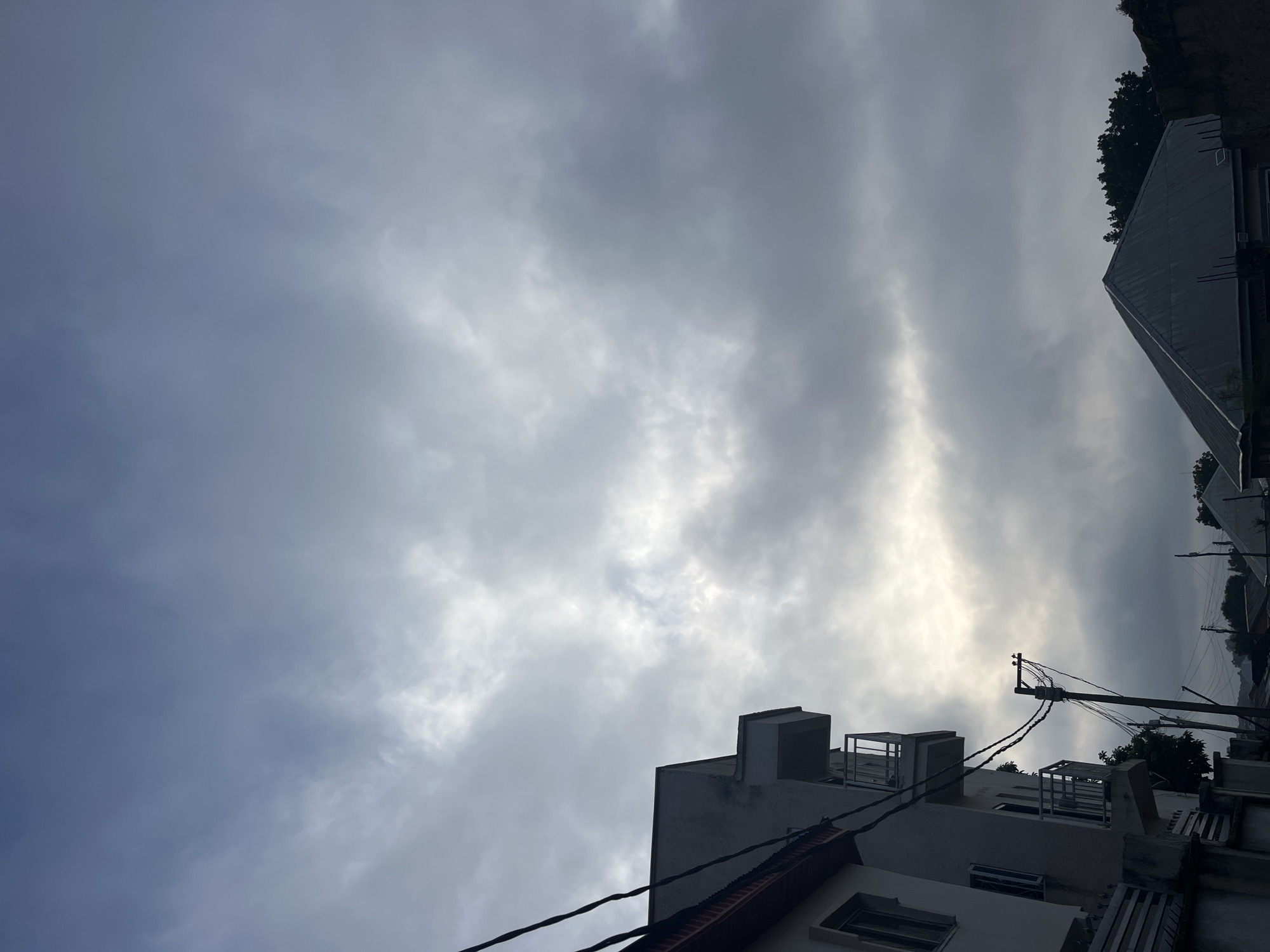 The image depicts a cloudy sky over a residential area, likely in Jos, Nigeria. The sky is dominated by grey and white clouds, with patches of brighter light suggesting the sun attempting to break through. In the foreground, the side of a multi-story building is visible, featuring balconies and windows. To the right, the angular roof of another building is prominent, partially obscured by trees. A utility pole with numerous wires stands between the buildings. The overall atmosphere is overcast and somewhat subdued.