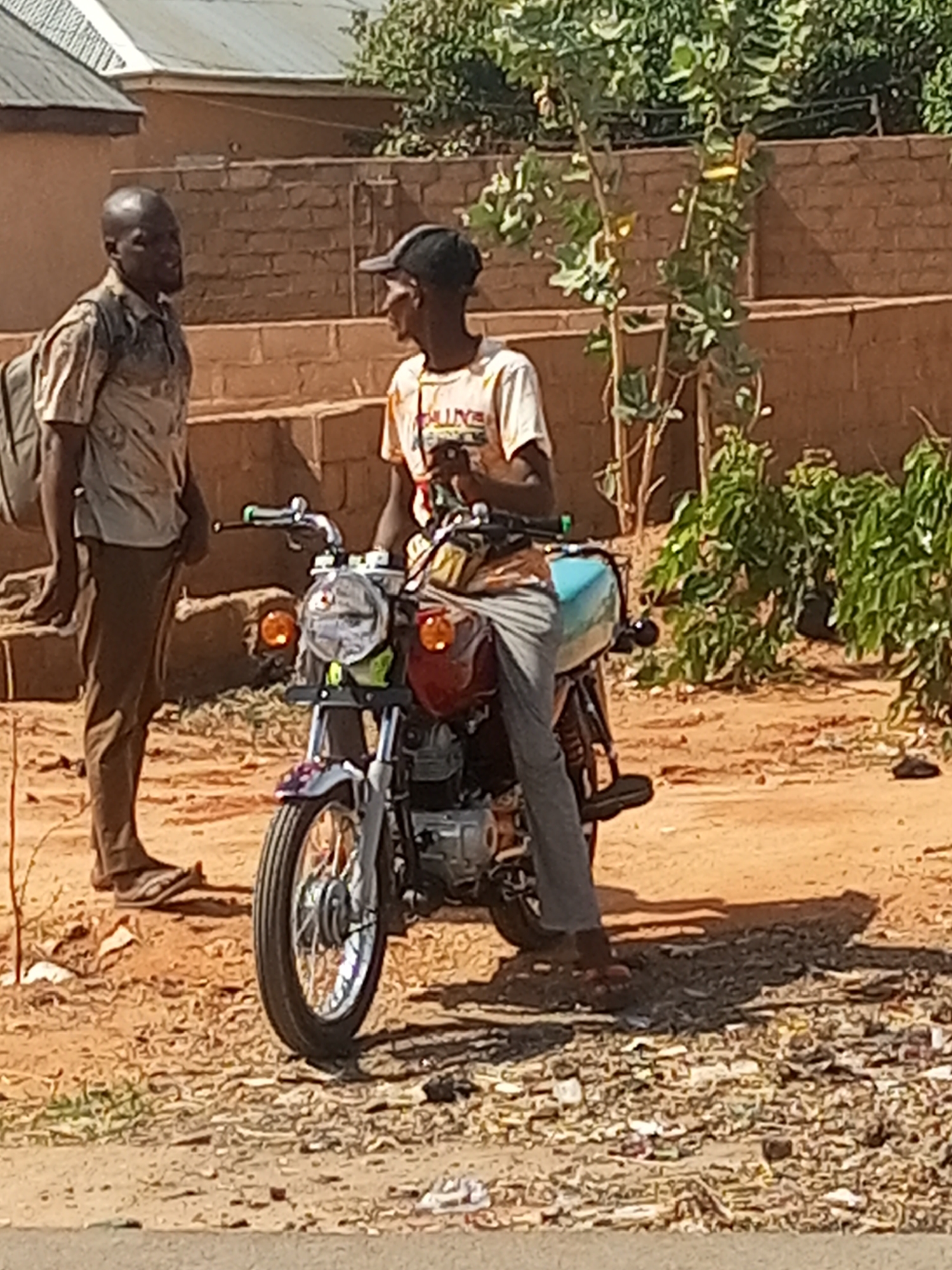 Two men are conversing next to a motorcycle in an outdoor, arid environment. The man on the left is standing, wearing a collared shirt, brown pants, and sandals. He carries a backpack on his back. To his right, another man is sitting on a motorcycle. He is wearing a light-colored T-shirt, grey pants, a black baseball cap, and sandals. The motorcycle is light-colored with a red gas tank. The ground is dirt and littered with debris. In the background, there is a brick structure, plants, and parts of buildings. The location is in the city of Unknown, Unknown.
