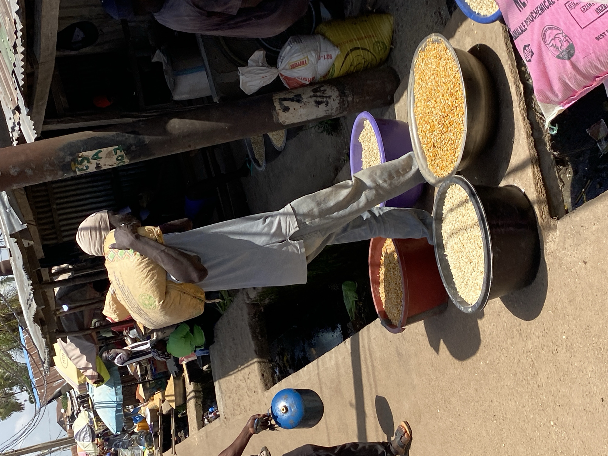 In Bukuru, Nigeria, a man is walking with a large sack of grain on his shoulder, moving from left to right across the frame. He is wearing a light-colored long-sleeved shirt, light-colored pants, and a head covering. To his left, another person, mostly out of frame, is holding a blue propane tank. In the foreground, there are several large containers of grain. A red tub is filled with yellow corn kernels, a black tub holds small white grains, and a purple tub contains more small white grains. In the background, a bustling market scene is visible with stalls and awnings. The ground is a textured, sandy surface.