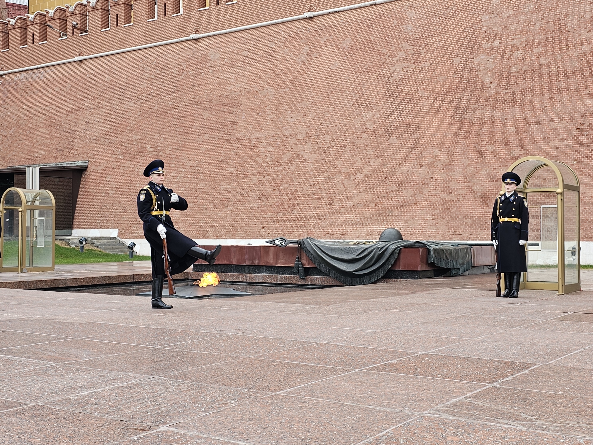 The image shows two honor guards at the Tomb of the Unknown Soldier in Moscow, Russia. The tomb is a dark stone platform with a bronze star in the center containing an eternal flame. One guard is standing at attention next to a small shelter. The other is performing a high-stepping goose-step while holding a rifle with a bayonet. The guards are wearing black, knee-length overcoats, black boots, and black hats with gold trim. Behind the guards is a high brick wall, part of the Kremlin. The ground is made of large square red granite tiles.