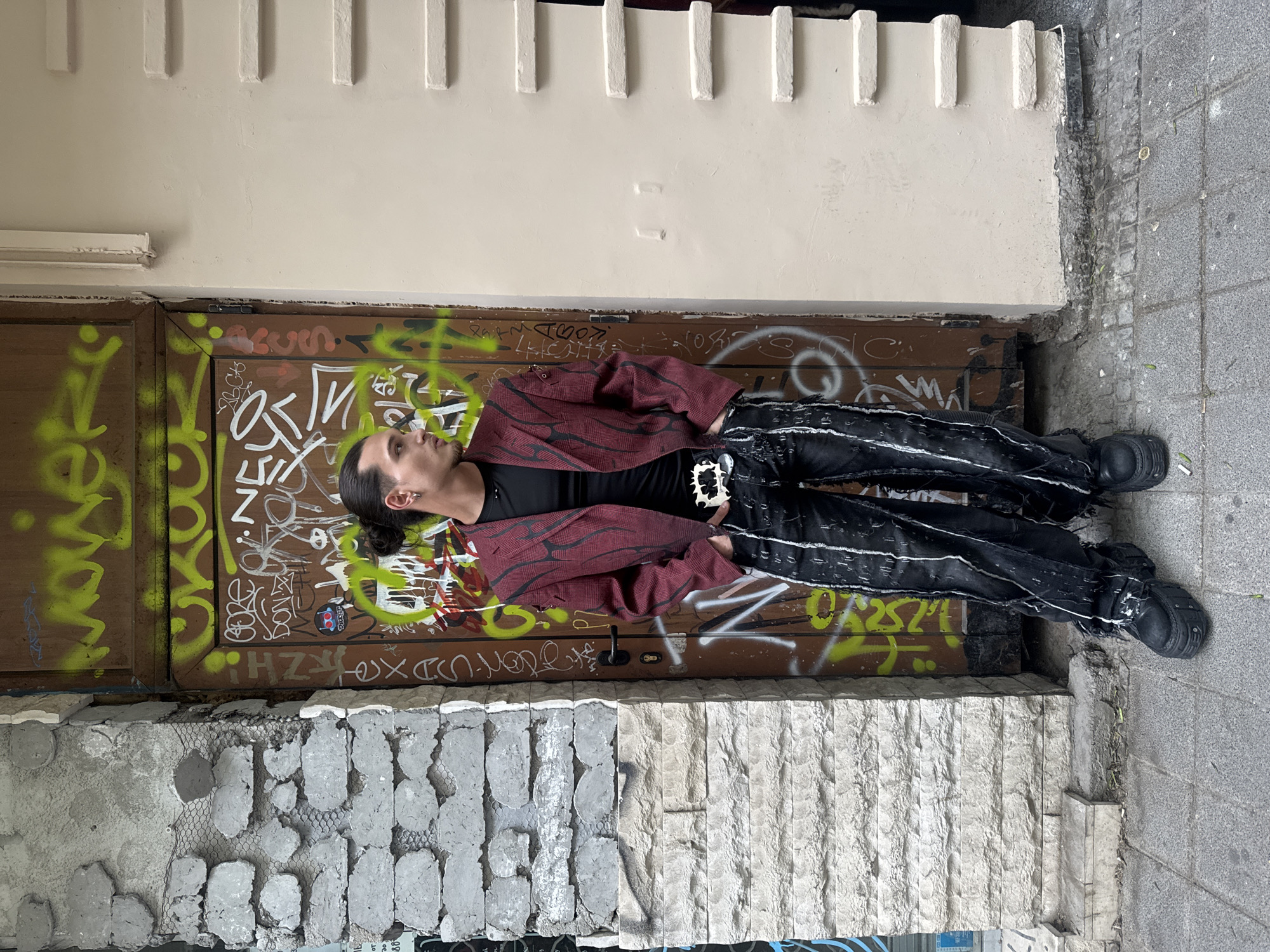 A person lies on a graffiti-covered brown door beneath a white-painted wall in Sofia, Bulgaria. The individual wears black pants with white stripes, a black shirt, and a maroon jacket. The person is wearing chunky black boots with prominent soles. The person has dark hair pulled back in a ponytail and is facing to the left of the frame. The door is adorned with green and white graffiti. Below the door is a section of wall featuring grey stone work. The ground is paved with grey cobblestones. A white wall with raised, evenly-spaced rectangular features runs parallel to the door. A portion of the wall has a creamy, white finish.