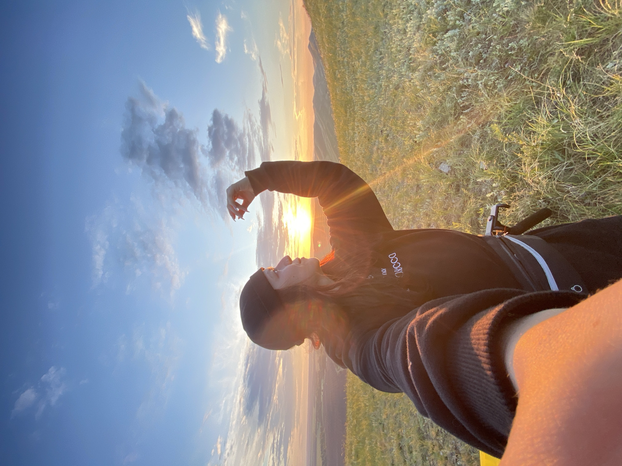 A young woman, wearing a black beanie and a dark hoodie with white lettering, stands on a grassy hill at sunset. Her right hand is raised, with her fingers extended, and she appears to be interacting with the light. The sun is low on the horizon, casting a warm orange glow across the landscape and creating lens flare. Rolling hills stretch into the distance, and the sky is a mix of blue and soft, scattered clouds. The image is a selfie, taken from a low angle, capturing the woman's profile and part of her arm in the foreground. The location is the vicinity of Абзаново, Russia, characterized by its natural, hilly terrain and open skies.