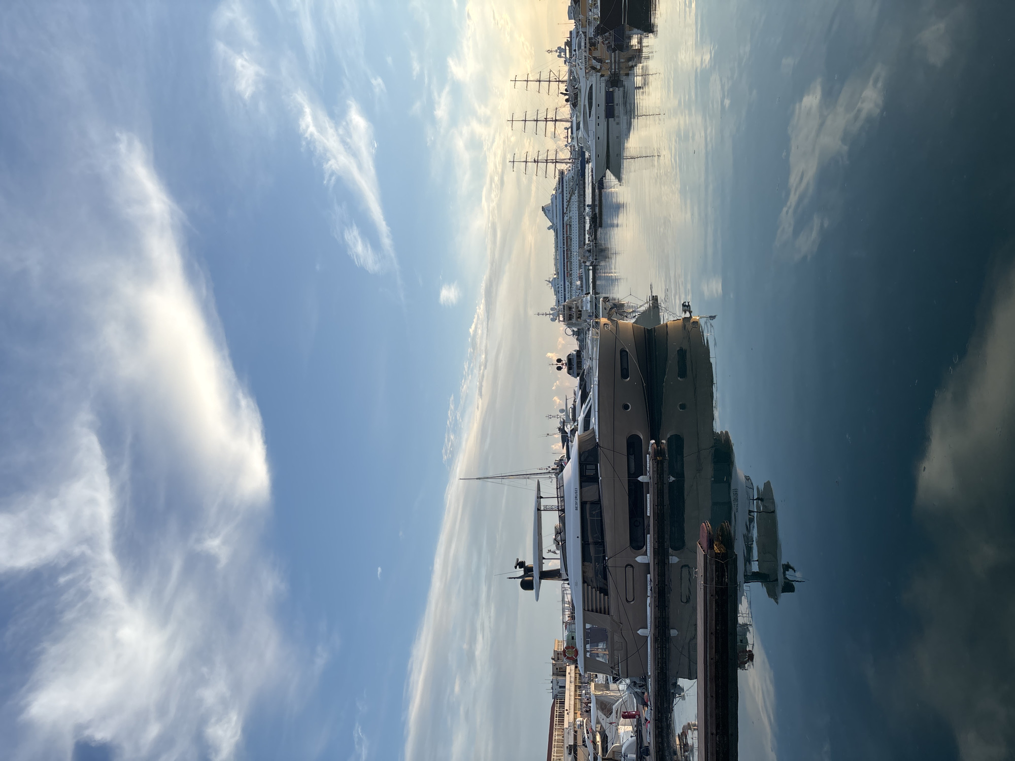 This is a vertical, eye-level, full shot of a marina in Sochi, Russia, with several yachts and a large cruise ship docked. The sky is a pale blue with wispy white clouds, and the water is calm and reflective, mirroring the sky and the boats. The most prominent yacht is in the foreground, a tan motor yacht with a white roof and dark tinted windows. It is docked alongside a wooden pier. In the background, a large white cruise ship is visible, along with several other yachts and sailboats, including some with tall masts. The sun appears to be low on the horizon, casting a warm glow on the scene and creating reflections on the water. The overall impression is one of tranquility and luxury.