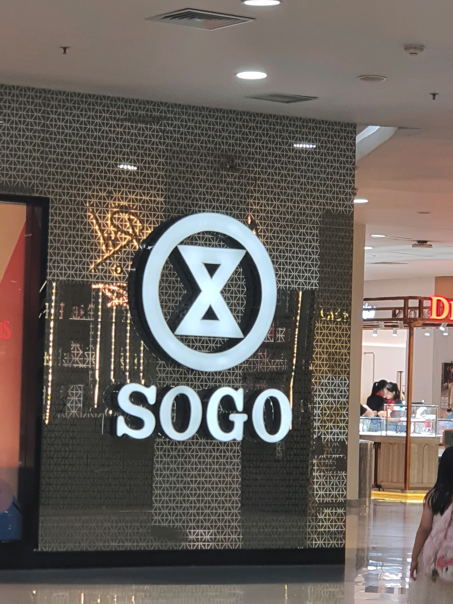 A sign with the word "SOGO" and a circular logo with an hourglass shape inside is mounted on a patterned wall. The sign is illuminated, and the logo is white. The wall has a repetitive geometric pattern in dark and light tones. To the right of the SOGO sign, there is a shop interior with display cases and two individuals. Further to the right, a person with dark hair wearing a light-colored dress is walking away from the camera. The scene appears to be inside a shopping mall, likely in North Jakarta, Indonesia, given the "SOGO" department store branding, which is a prominent retailer in the region. The ceiling has recessed lighting and vents.