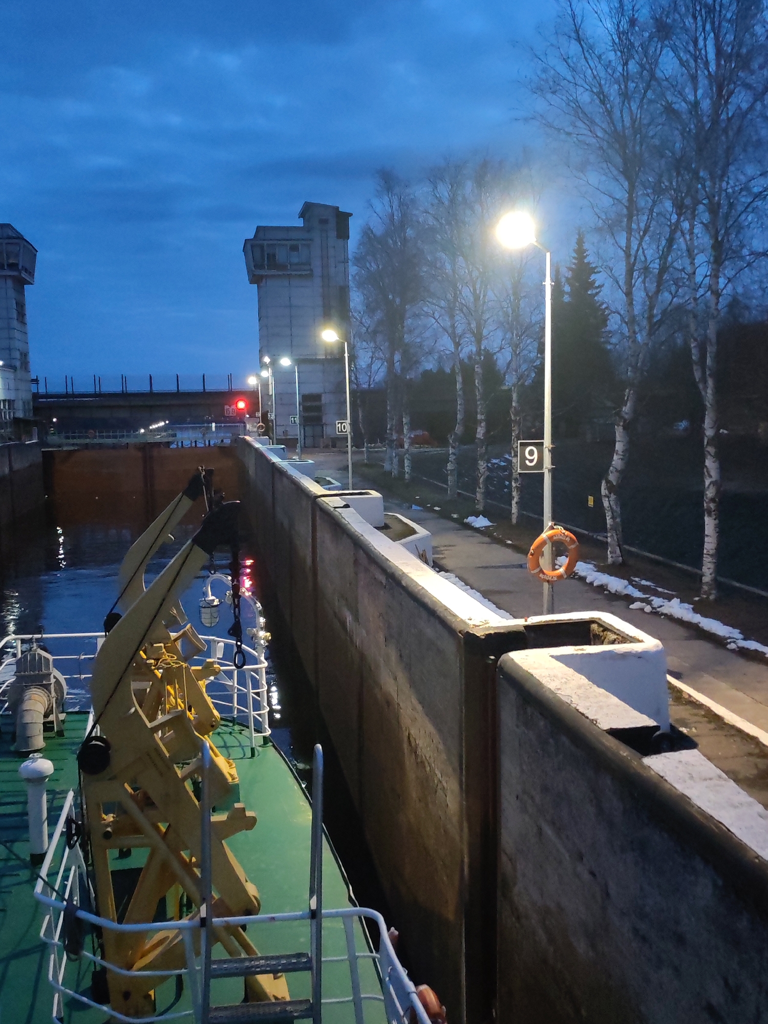 A low-light, wide-angle shot depicts a ship inside a canal lock system at dusk or night. The sky is a dark, cloudy blue, illuminated by streetlights and the ship's own lights, which reflect on the water.
In the foreground, the green deck and white railings of a ship are visible, along with prominent yellow crane-like equipment on its bow. The vessel is situated next to a tall, dark concrete lock wall. Along the path beside the wall, patches of snow or ice are visible on the ground, indicating cold weather. An orange lifebuoy hangs from a lamppost that displays the number "9" on a white sign. Other lampposts further along the path also bear numerical signs, including "10," "11," and "12."
The lock chamber's dark water is visible ahead of the ship, with a large lock gate partially seen beyond. A bright red signal light is prominently displayed in the distance, likely indicating a stop or closed gate. In the background, two imposing concrete control towers with multiple windows flank the lock entrance, connected by a bridge-like structure. Bare deciduous trees, with some evergreens, line the distant banks. No individuals are visible in the scene. The overall impression is one of a quiet, industrial setting in cold, dim conditions.