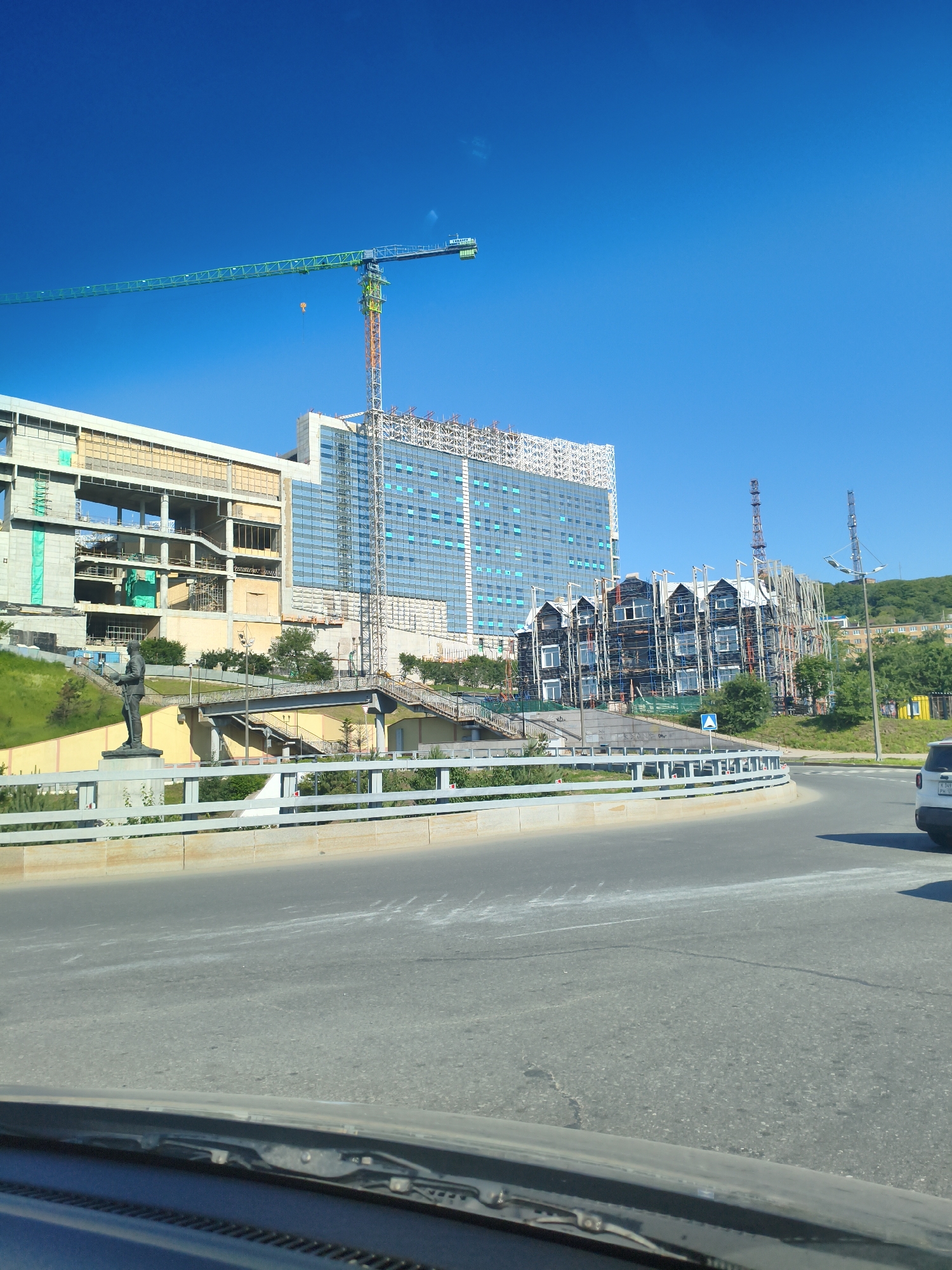 A wide-angle, eye-level, outdoor, daytime, vertical shot captures a construction site with multiple buildings under development in Vladivostok, Russia. In the foreground, a curved asphalt road is visible, with a light-colored concrete barrier separating it from a green, grassy slope. A statue of a standing man in uniform is positioned on the slope, facing left, with a ramp or bridge structure behind it.
Further back, a large, multi-story building with a blue glass facade is under construction. To its left, another building is in an earlier stage of construction, with exposed concrete and scaffolding. To the right of the blue-glass building, a smaller, dark-colored building with a series of gabled roofs and numerous windows is also under construction, with scaffolding covering its exterior.
Dominating the sky, a long, green construction crane extends horizontally across the frame, with its vertical support structure visible. Two other tower cranes are also present in the background. The sky is a clear, bright blue. The overall scene depicts a developing urban area with ongoing construction projects.