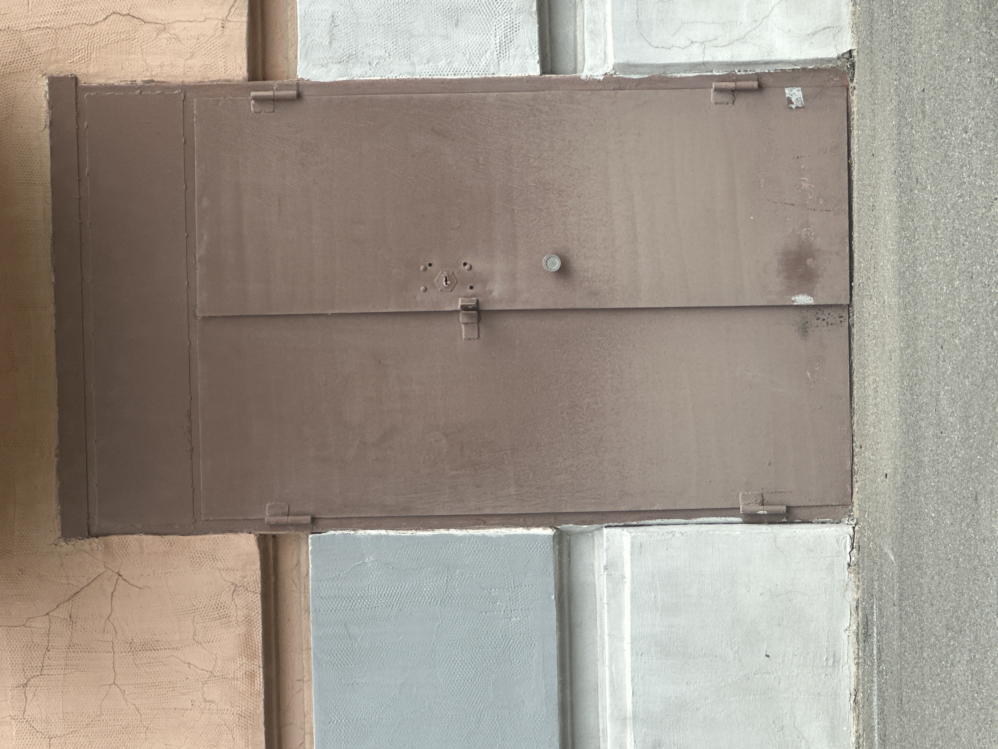 The image depicts a closed, double-doored, brown metal garage or service entrance on the ground floor of a building in Saint Petersburg, Russia. The doors are robust and appear weathered, with visible hinges on the right side of the right door and the left side of the left door. A rectangular bolt lock is mounted on the left door, just below a keyhole and what appears to be a circular knob or handle. The wall surrounding the doors is painted in distinct horizontal sections: a light peach or salmon color with visible cracks at the top, a muted blue-grey textured section below that, and a light grey textured section at the bottom, just above the asphalt ground. The asphalt extends into the foreground, suggesting a street or driveway. The overall impression is one of utilitarian architecture, common in urban environments. There are no people or animals present, and no actions are being taken. The scene is static and focuses solely on the architectural elements.