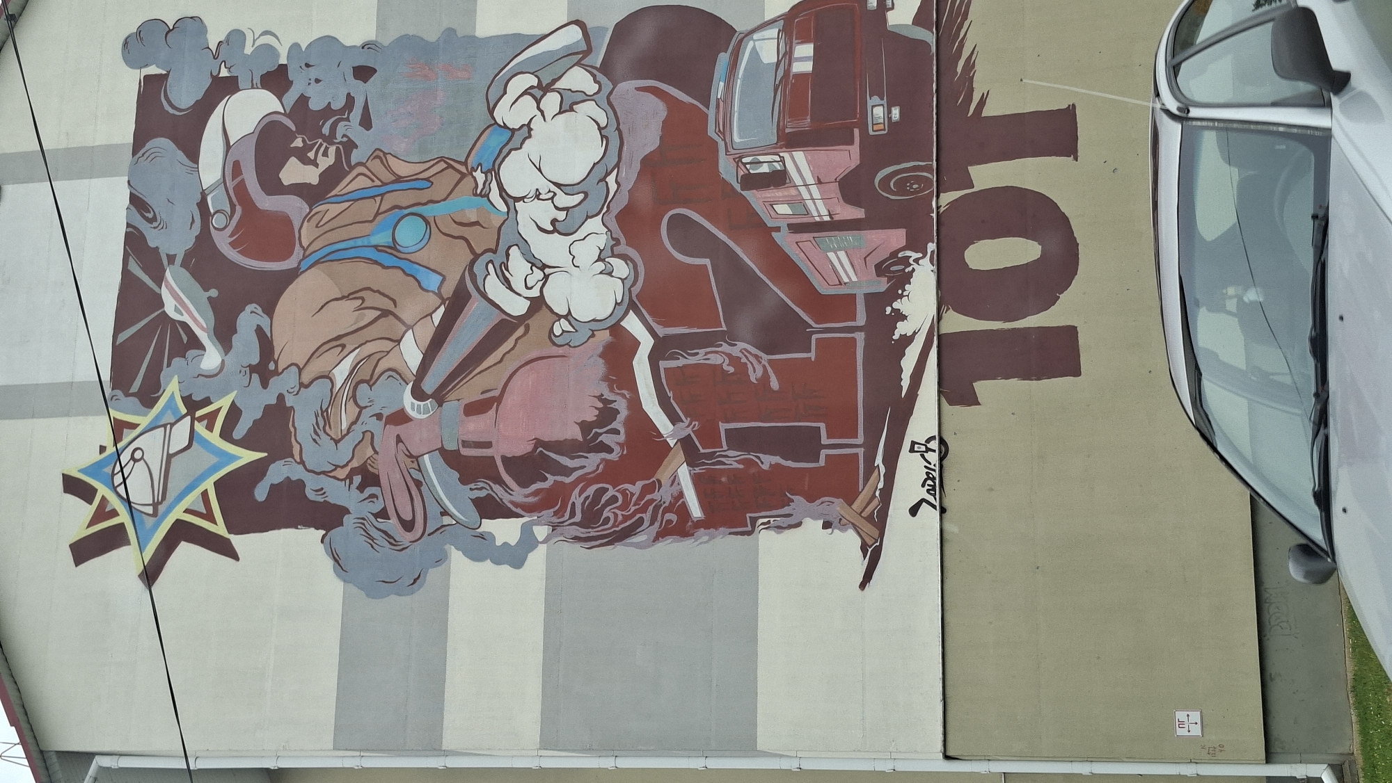 This is a photograph of a mural painted on the side of a building in Myadzyel, Belarus. The mural depicts a firefighter in full gear, holding a hose and surrounded by smoke and flames. Behind the firefighter are the numbers "112", representing the European emergency number. A red fire truck is also visible in the mural. Below the "112", the number "101" is painted, which is the emergency number for firefighters in Belarus. A silver car is parked in front of the building, partially obscuring the lower portion of the mural. The building itself appears to be a multi-story residential structure, with horizontal grey and cream-colored stripes on its facade. There is a white electrical conduit running vertically along the left side of the building. A small white sign with black markings is attached to the wall near the conduit. The sky is visible at the top of the image, and the scene is illuminated by daylight.