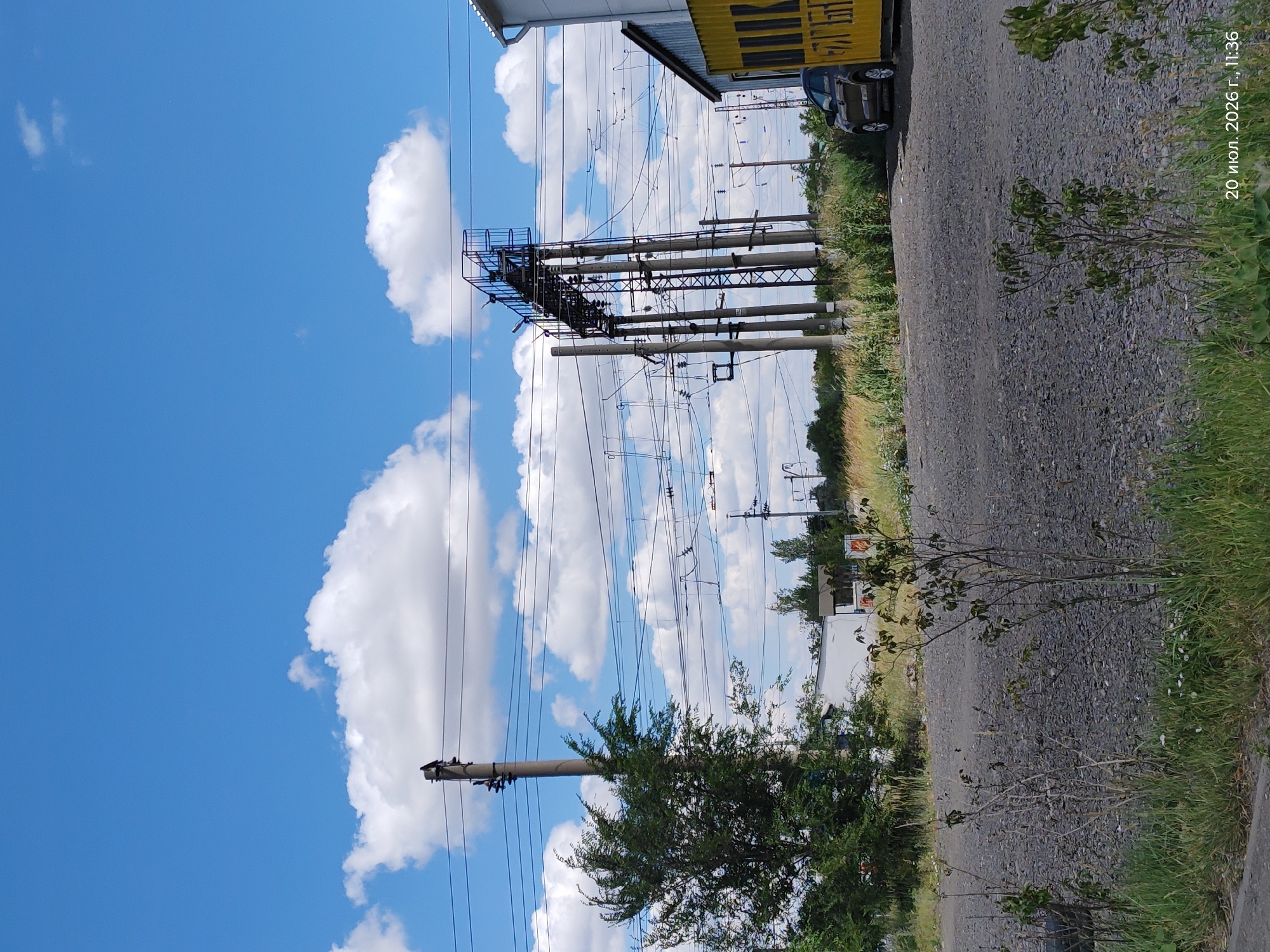 A medium-shot, eye-level, outdoor, daytime, horizontal image shows a view of overhead railway power lines and supporting poles. In the foreground, there is a gravel-covered area with some sparse green vegetation and dried brush. A dark-colored car is parked on the right side of the frame next to a yellow shipping container with black lettering. In the background, there are trees and other structures, including a white building with a small windowed structure attached. The sky is blue with scattered white cumulus clouds. The image has a timestamp in the lower right corner reading "20 июл. 2026 г., 11:36". The location is identified as Astana, Kazakhstan.