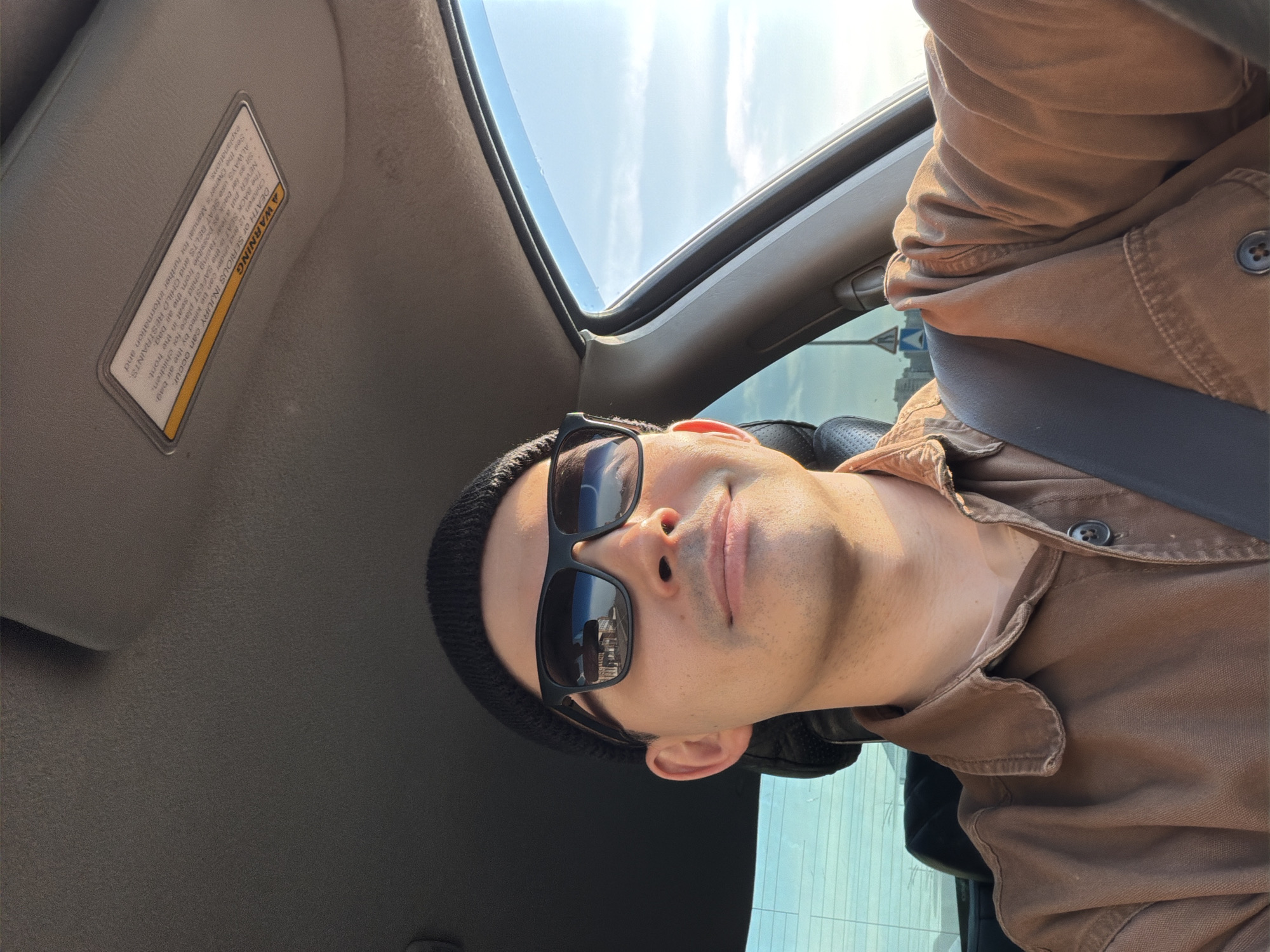 The image shows a man wearing a black beanie and sunglasses, sitting in a car. He is wearing a brown shirt and a seatbelt. Part of the car interior is visible, including the sun visor with a warning label. The car appears to have a sunroof. The reflection in the sunglasses suggests a city scene with buildings. It is not possible to identify the specific location of Izhevsk, Russia.