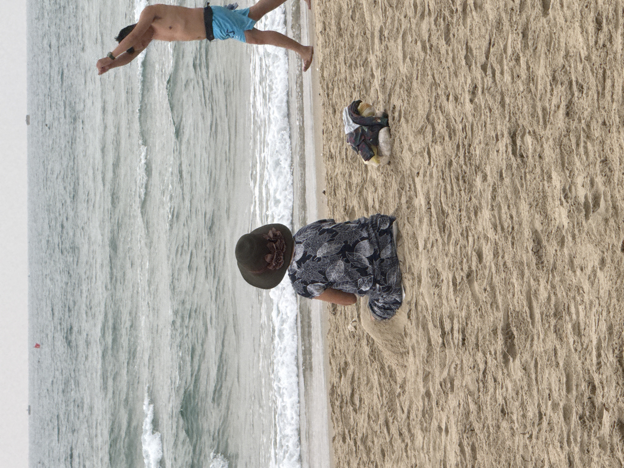 A beach scene is presented. A man wearing blue swimming trunks is diving into the ocean. A woman wearing a dark hat and a leaf-printed shirt stands on the sand near the water’s edge. A pile of clothes is located on the sand between them. The location is likely My Khe beach in Đà Nẵng, Vietnam.
