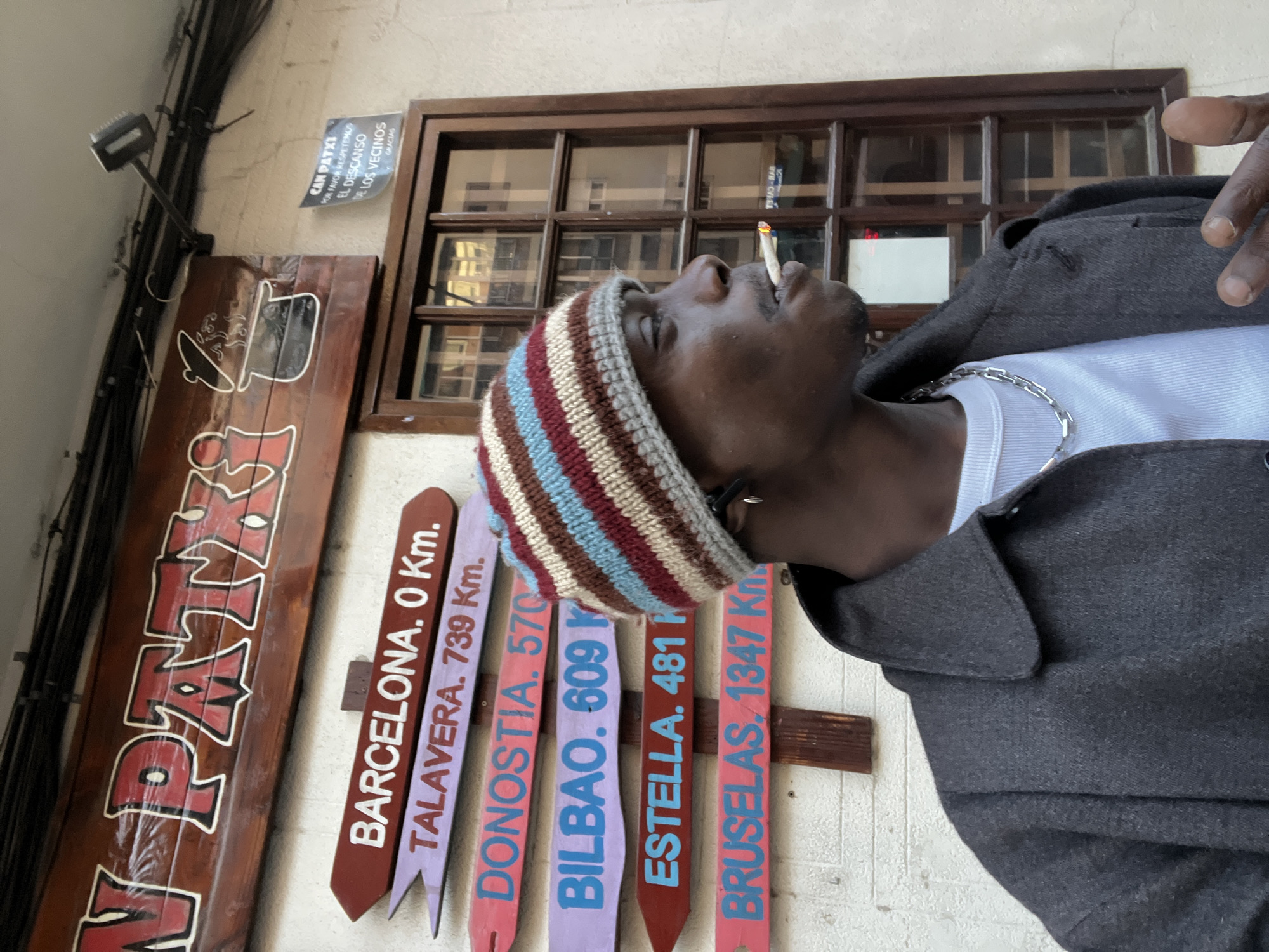 A man wearing a striped beanie, silver chain, white T-shirt, and dark gray wool coat is smoking a cigarette in an outdoor setting. He is positioned near a wooden sign that reads "J-PATXI." To the left of the man are multiple directional signs indicating distances to various cities, including "BARCELONA. 0 Km.," "TALAVERA. 739 Km.," "DONOSTIA. 570 Km.," "BILBAO. 609 Km.," "ESTELLA. 481 Km.," and "BRUSELAS. 1347 Km." A sign above the wooden sign indicates "CAN PATXI" and a request to respect the rest of the neighbors in Spanish. The location is likely near the directional sign indicating the proximity to Barcelona. The image is taken at an upward angle.