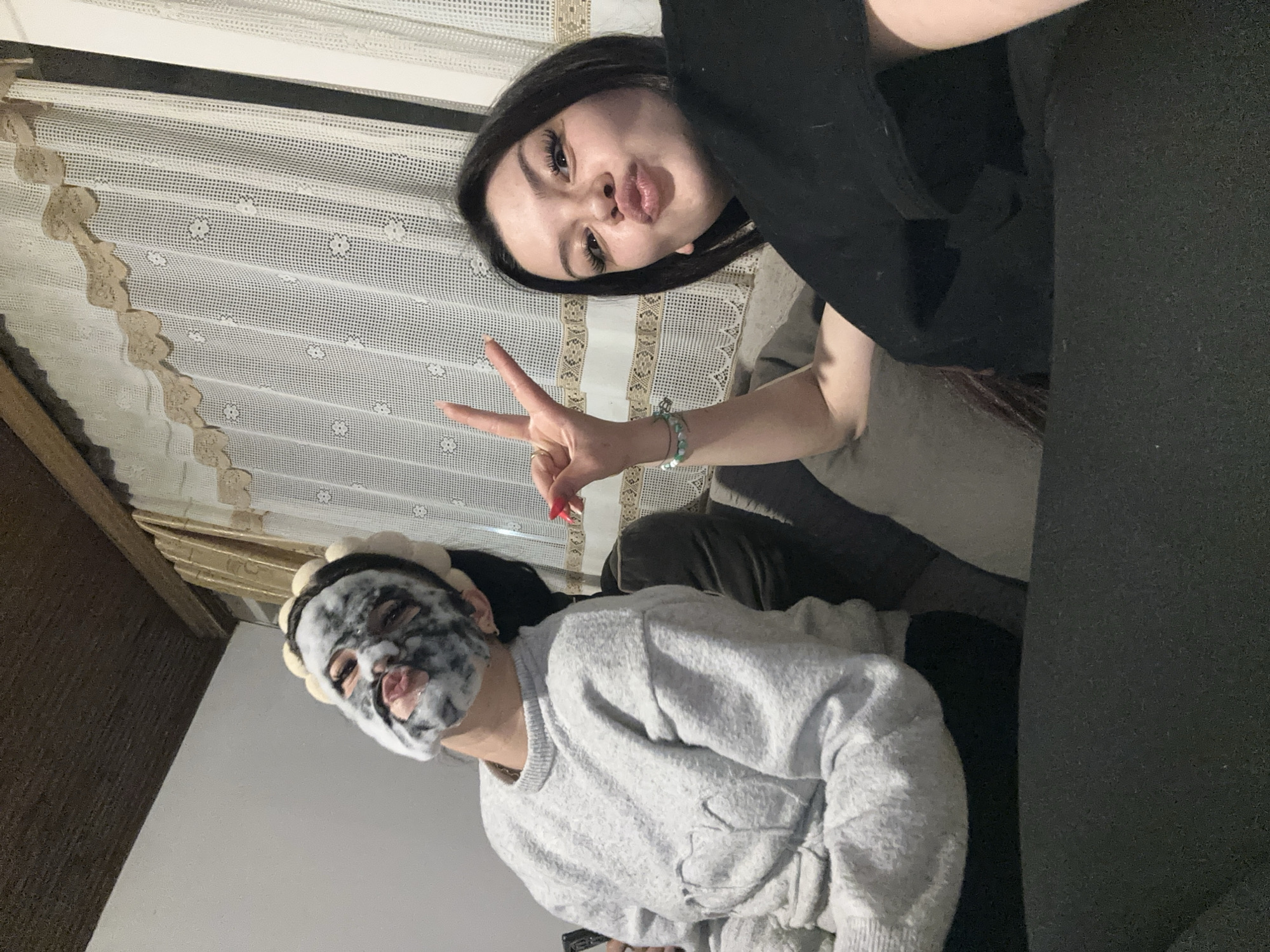 Two females are in a room in Chubra, Bulgaria. One is wearing a grey sweater and a foamy face mask. The other has dark hair and is wearing a black shirt. She is making a peace sign with her right hand. There is a patterned curtain in the background.