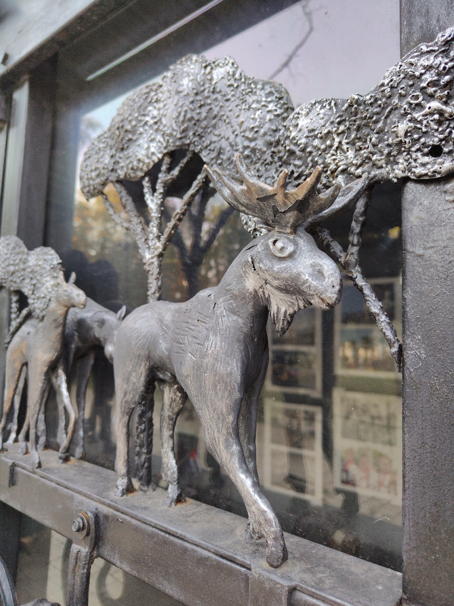 The image is a close-up, low-angle shot of a metal sculpture of moose, likely part of a decorative fence or gate. The sculpture is grey and appears to be made of wrought iron or a similar metal. In the foreground, a large moose is depicted in profile, facing left. It has detailed antlers, a textured coat, and its legs are positioned as if walking. Behind this moose, to the left, are two smaller moose, also in profile, one partially obscuring the other. These appear to be a mother and calf. The sculpture is set against a backdrop of a reflective surface, possibly glass, which shows a blurred reflection of trees and possibly an interior space with framed items. The metalwork of the sculpture itself is highly textured, with a rough, almost organic surface, especially on the trees depicted in the background of the sculpture. The foreground also shows a metal railing with a bolt and hinge mechanism, indicating it is part of a larger structure. There are no people visible in the image. The environment appears to be outdoors, given the reflection of trees. This particular sculpture is located at the entrance to the Donetsk Zoo in Donetsk, Ukraine.