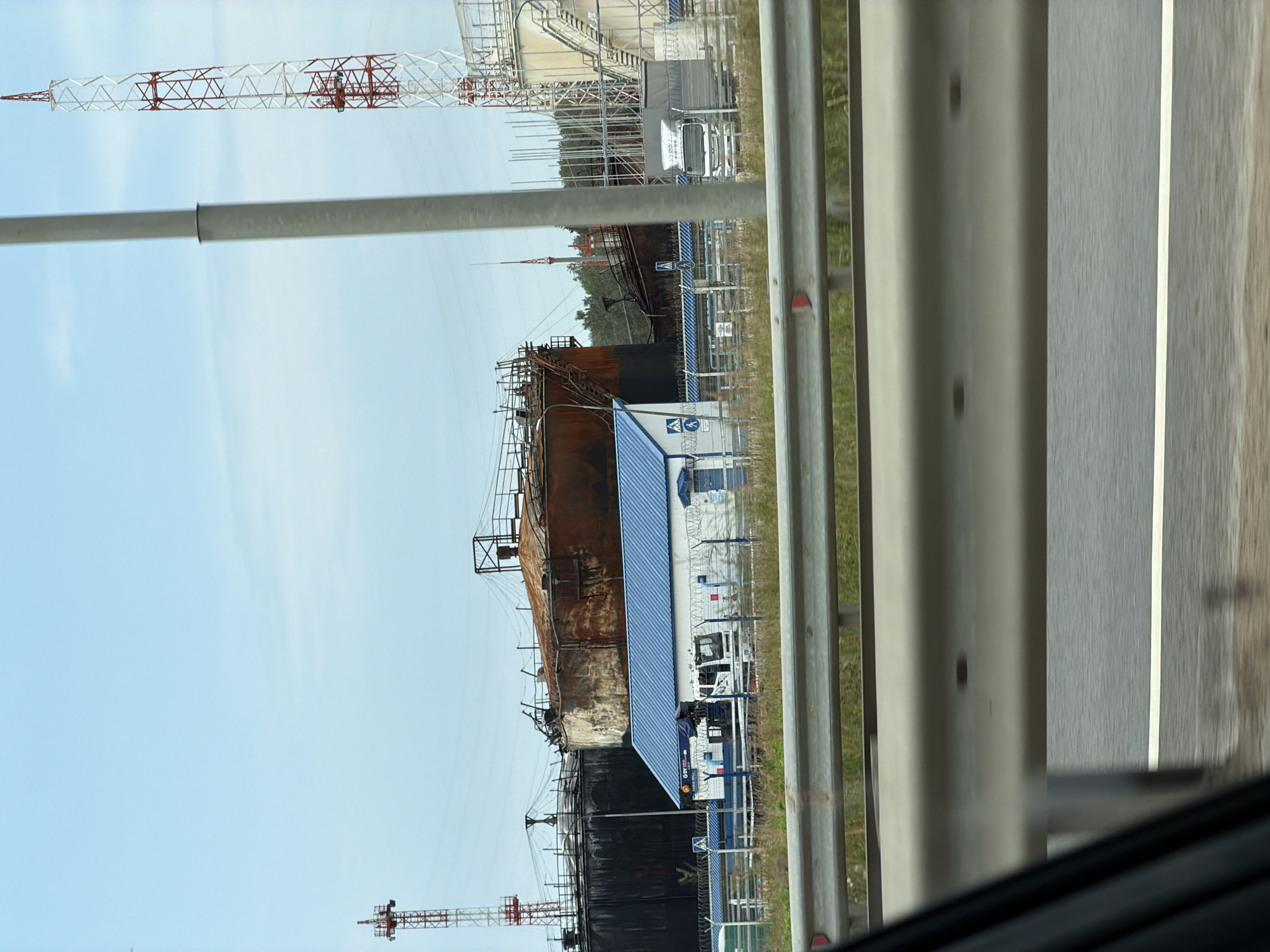 The image depicts an industrial area in Radishchevo, Russia, characterized by large, rusty oil storage tanks and associated infrastructure. In the foreground, a grey guardrail runs horizontally across the frame, with a road surface visible below it. Behind the guardrail, a grassy area is present. The main focus of the image is a complex of industrial structures. Two prominent, large, cylindrical oil storage tanks are visible, one appearing heavily rusted and discolored, suggesting age or damage, and the other darker, possibly blackened. A smaller, white building with a blue roof is situated in front of the tanks, with a fence surrounding it. Several vehicles are present within the fenced area, including a white truck and what appears to be a white utility vehicle or small truck. Two red and white transmission towers are visible in the background, one to the left and another taller one to the right, reaching towards the light blue sky. The overall environment suggests an operational or formerly operational industrial site. There are no visible people in the image.