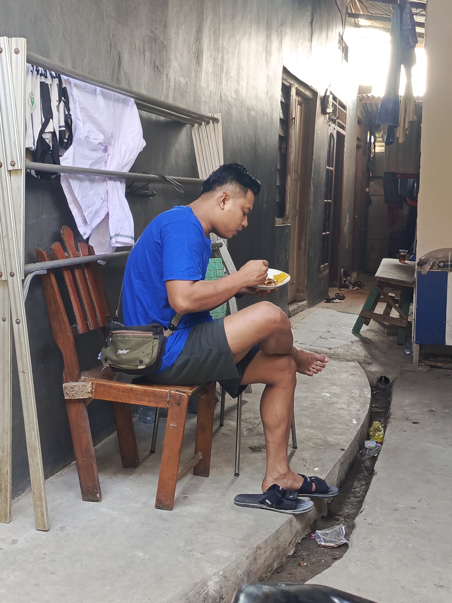 A man is seated on a wooden chair in an alleyway in Tangerang, Indonesia, eating from a plate. He is wearing a blue t-shirt, dark shorts, and black sandals. A brown and green satchel is slung across his body. Clothes are hanging on a rack to his left, and more clothes and a small table with a glass of liquid are visible in the background. The alleyway has a concrete floor with a drainage ditch running along its side, containing some debris. The walls of the buildings are textured and appear to be painted. The lighting suggests it is daytime, with sunlight filtering in from the right side of the frame.