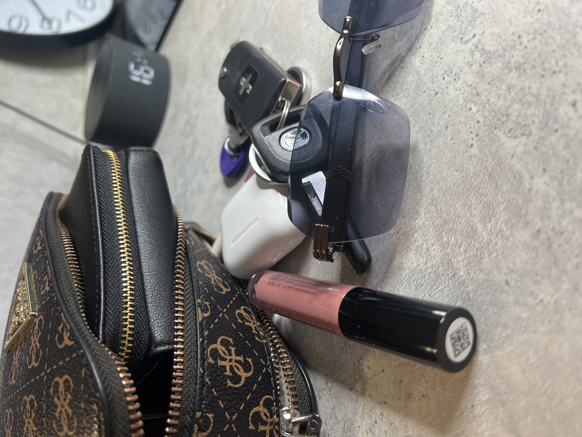 The image captures a collection of personal items laid out on a light grey, textured surface, possibly a countertop or table. The arrangement appears casual, suggesting the items have just been placed down or emptied from a bag.
In the foreground, a brown and black patterned purse with prominent gold zippers is visible. The purse is adorned with a repeated "GUESS" logo pattern and features a metallic, triangular "GUESS" emblem on its front flap. Its zippers are partially open, offering a glimpse into the black interior.
To the right of the purse, a cluster of everyday essentials is arranged. A set of keys, including a car remote key fob and another key bearing a visible "TOYOTA" logo, are held together on a keyring that also features a small purple tag. Partially beneath the keys and adjacent to the purse, a white AirPods charging case is visible.
A pair of dark-tinted sunglasses with a metallic bridge and hinges rests on top of some of the keys. Below the sunglasses, a tube of lip gloss or liquid lipstick, with a black body and a clear section showcasing a pinkish-nude shade, is laid horizontally. A small QR code is clearly visible on the bottom end of its cap.
In the blurred background, two timekeeping devices can be discerned. A black, cylindrical digital alarm clock or timer displays the time "1:31". Behind it, part of a round analog clock face with numerical markings is also visible.
The image does not depict any people or specific activity, focusing solely on these everyday objects. No location cues are discernible that specifically point to Tyumen, Russia, other than the general nature of the items.