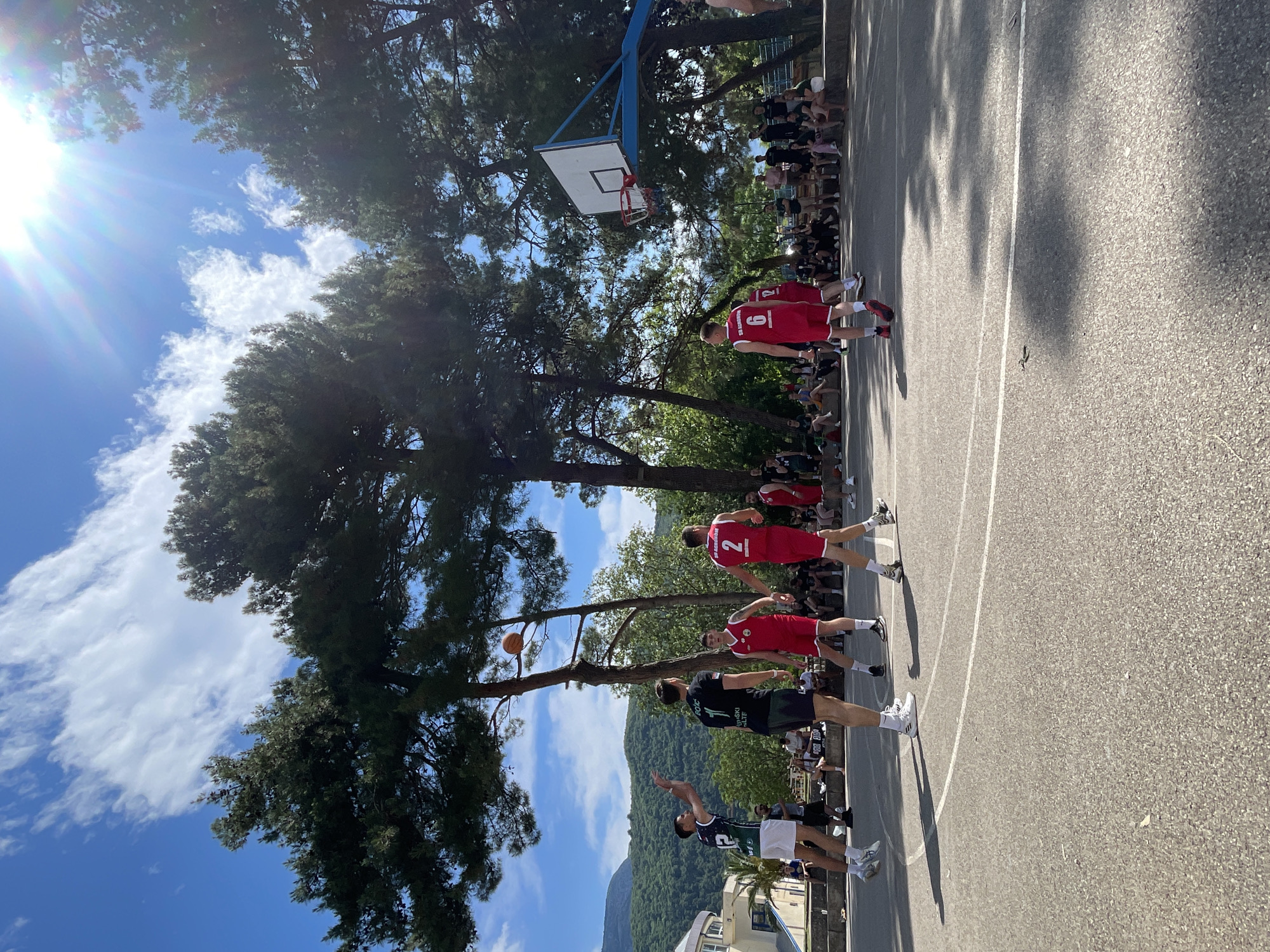 The image shows a basketball game being played outdoors on a court in the city of Unknown, Unknown. A group of people are watching the game as spectators. The players are wearing sports uniforms. There are several trees surrounding the court. The sky is bright blue with scattered clouds. A mountain range is visible in the background.