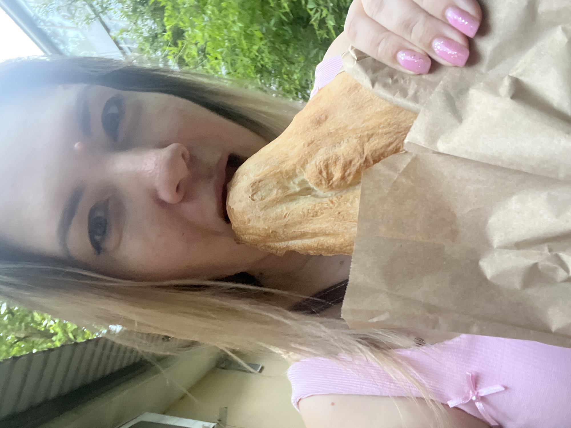 A woman is eating bread outside, possibly in Sochi, Russia. She is wearing a pink top with a bow detail. Her nails are painted a sparkly pink. The bread is wrapped in brown paper. In the background, there is greenery and a building.