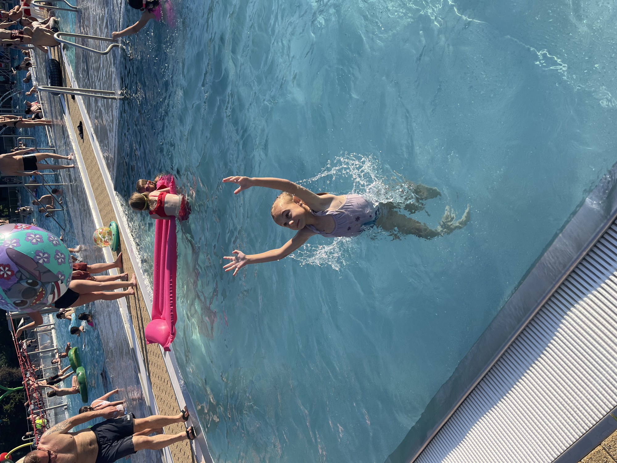 In this image, a group of people are enjoying a day at a swimming pool. The main focus is a young girl with blonde hair, wearing a purple swimsuit, as she swims through the bright blue water, creating a splash around her. Her arms are outstretched, and she appears to be having fun.
Several other individuals are present in and around the pool. Two children are near the edge of the pool, with one lying on a pink inflatable raft and the other partially submerged in the water. People are also seen standing and sitting along the pool's edge, some with swimsuits on, indicating a leisurely day of swimming and relaxation. A large inflatable ball with a Stitch character from Lilo & Stitch is visible near the pool's edge, suggesting a playful atmosphere.
The sun is shining brightly, casting shadows and illuminating the water, implying it is daytime. The setting appears to be an outdoor swimming pool, with a textured edge and a slatted walkway along the side. The water is clear and inviting, with ripples and splashes indicating activity. The overall mood seems to be joyful and carefree, typical of a summer day spent at the pool. No visible text is present in the image.